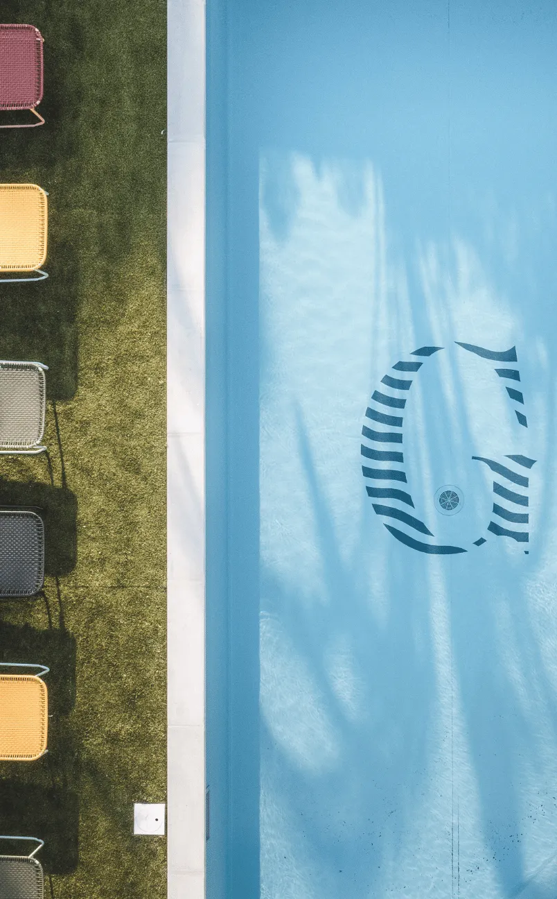 Veduta aerea di una piscina blu pulita con un logo stilizzato «G» nella parte inferiore e sedie colorate allineate sull'erba verde accanto.