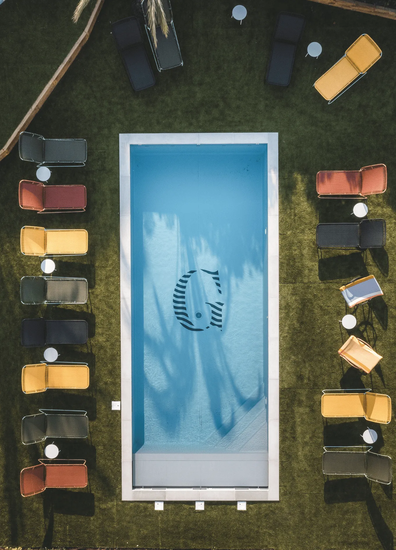 Vista dall'alto di una piscina rettangolare blu con un grande logo G nella parte inferiore, circondata da erba verde e sedie a sdraio colorate con tavolini rotondi.
