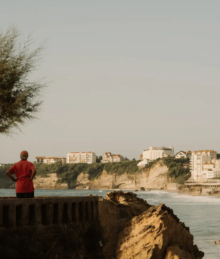 Person in red shirt and cap standing on stone railing overlooking rocky coastline with cliffs and buildings in the distance.