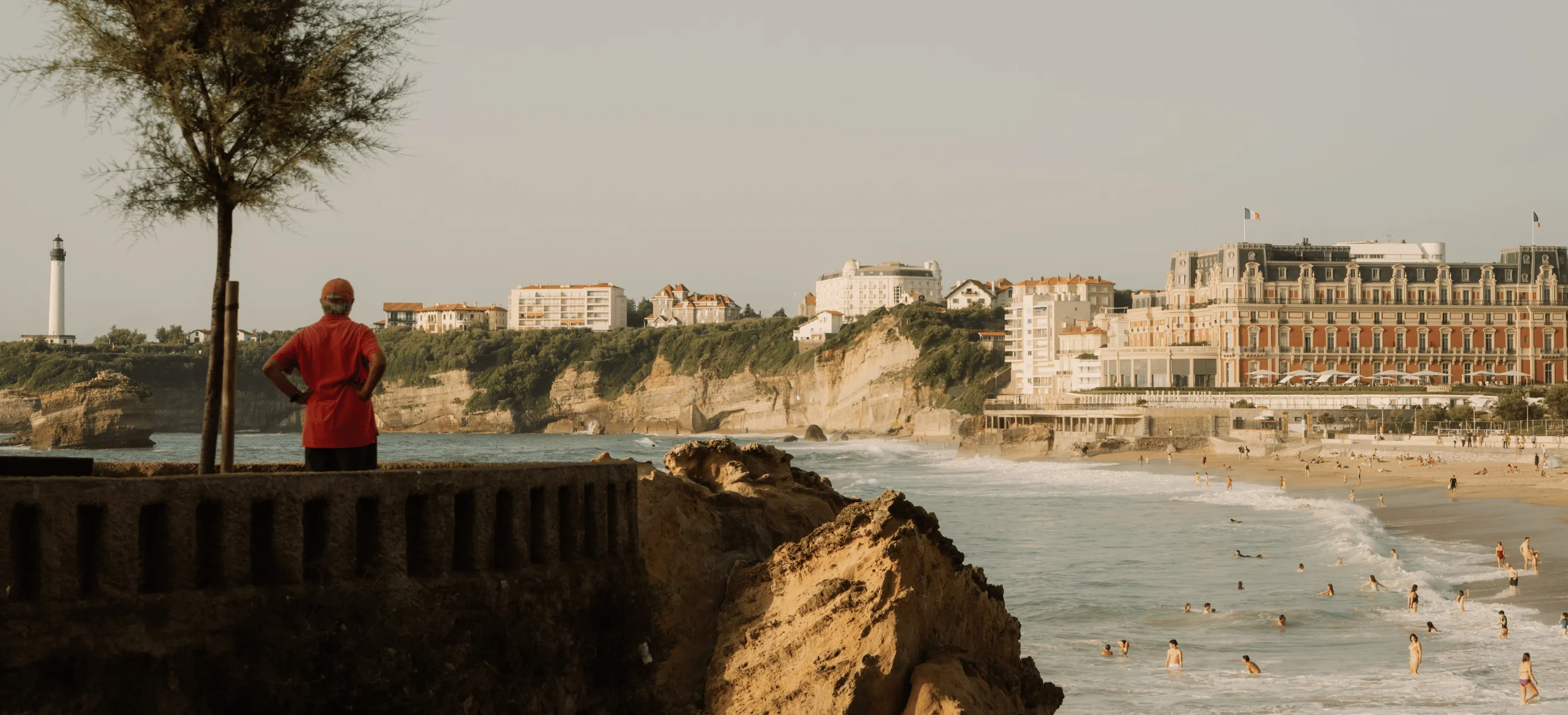 Person in red shirt standing by a stone railing overlooking a beach with swimmers, rocky cliffs, and buildings including a large hotel and a lighthouse in the distance.