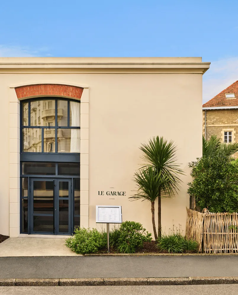 Facade of a building with a large arched window and door, a sign reading 'Le Garage,' and surrounding greenery.