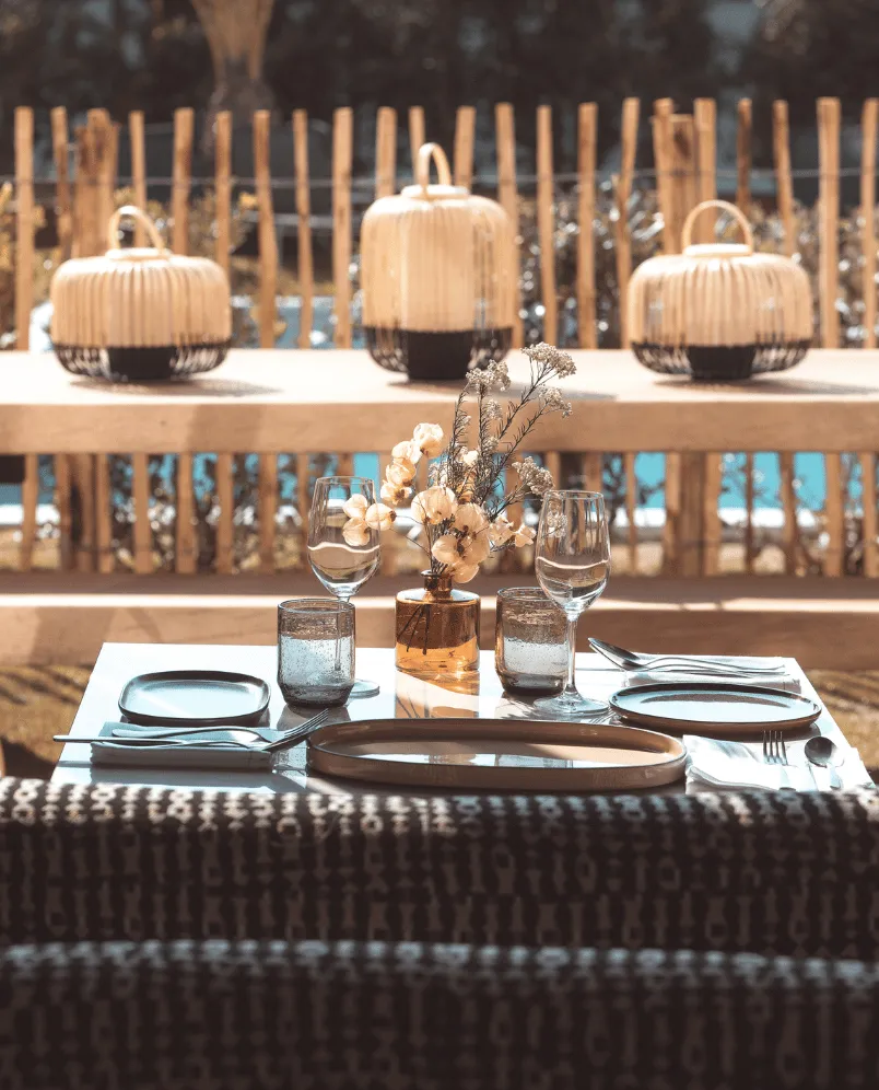 Outdoor dining table set for two with glassware, plates, cutlery, and a vase with dried flowers, with woven lanterns and a wooden fence in the background.