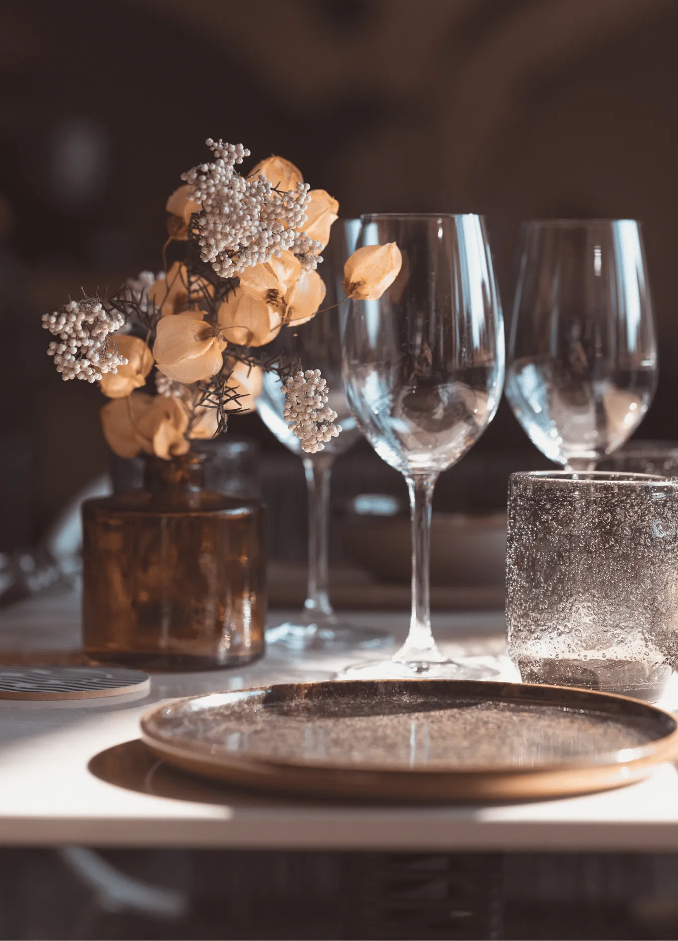 Close-up of elegant dining table setting with glassware, a textured plate, and dried flowers in a brown glass vase.