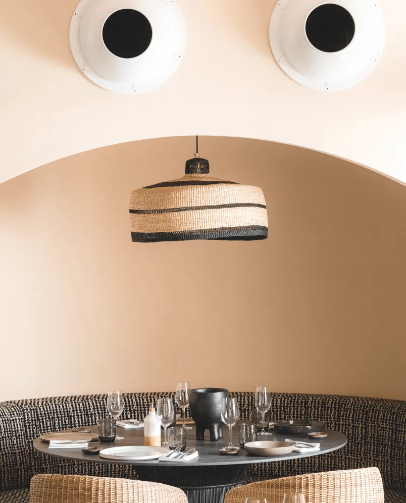 Elegant restaurant dining area with a round table set with glassware, plates, and napkins, wicker chairs, and a striped pendant light under a beige arch.