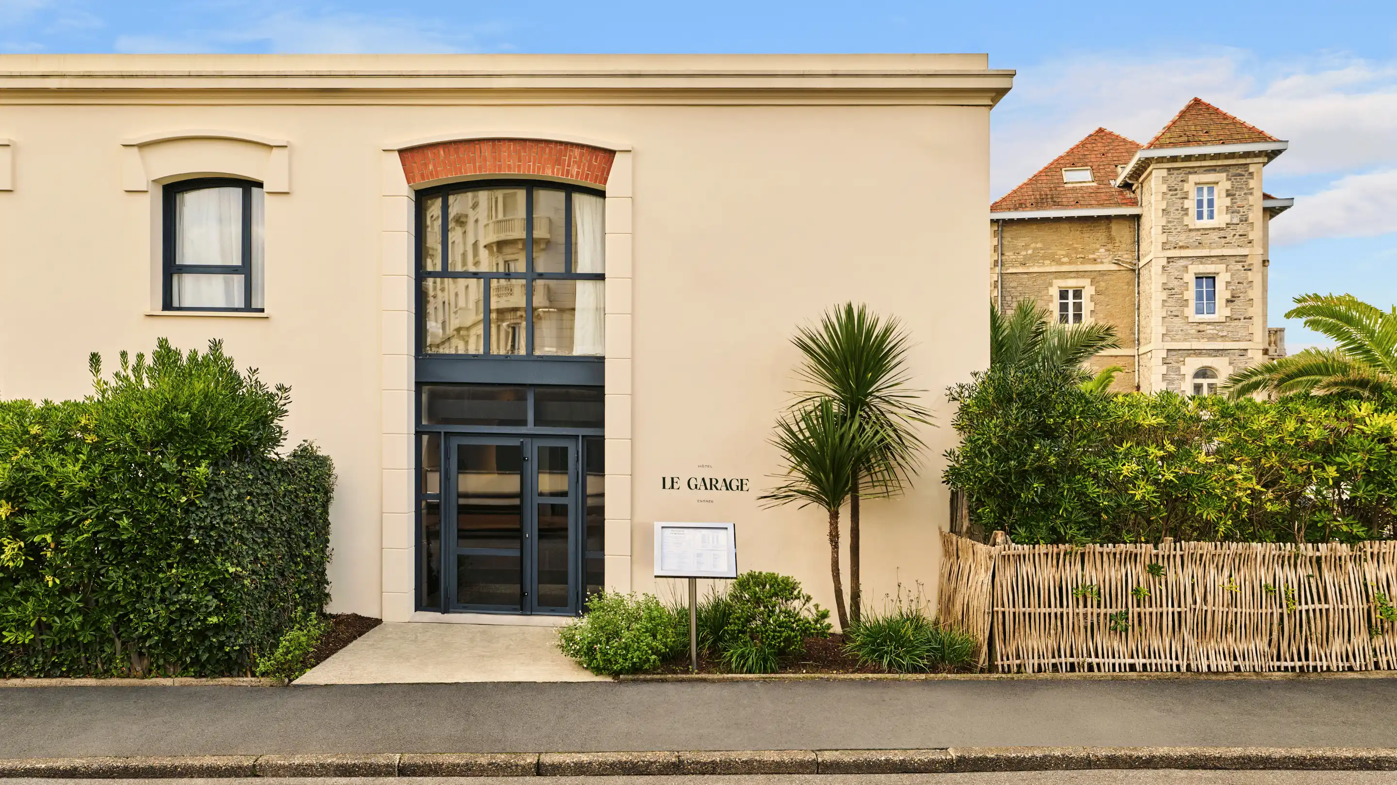 Entrance of Le Garage Hotel with large black glass door and window, surrounded by green bushes and palm trees.