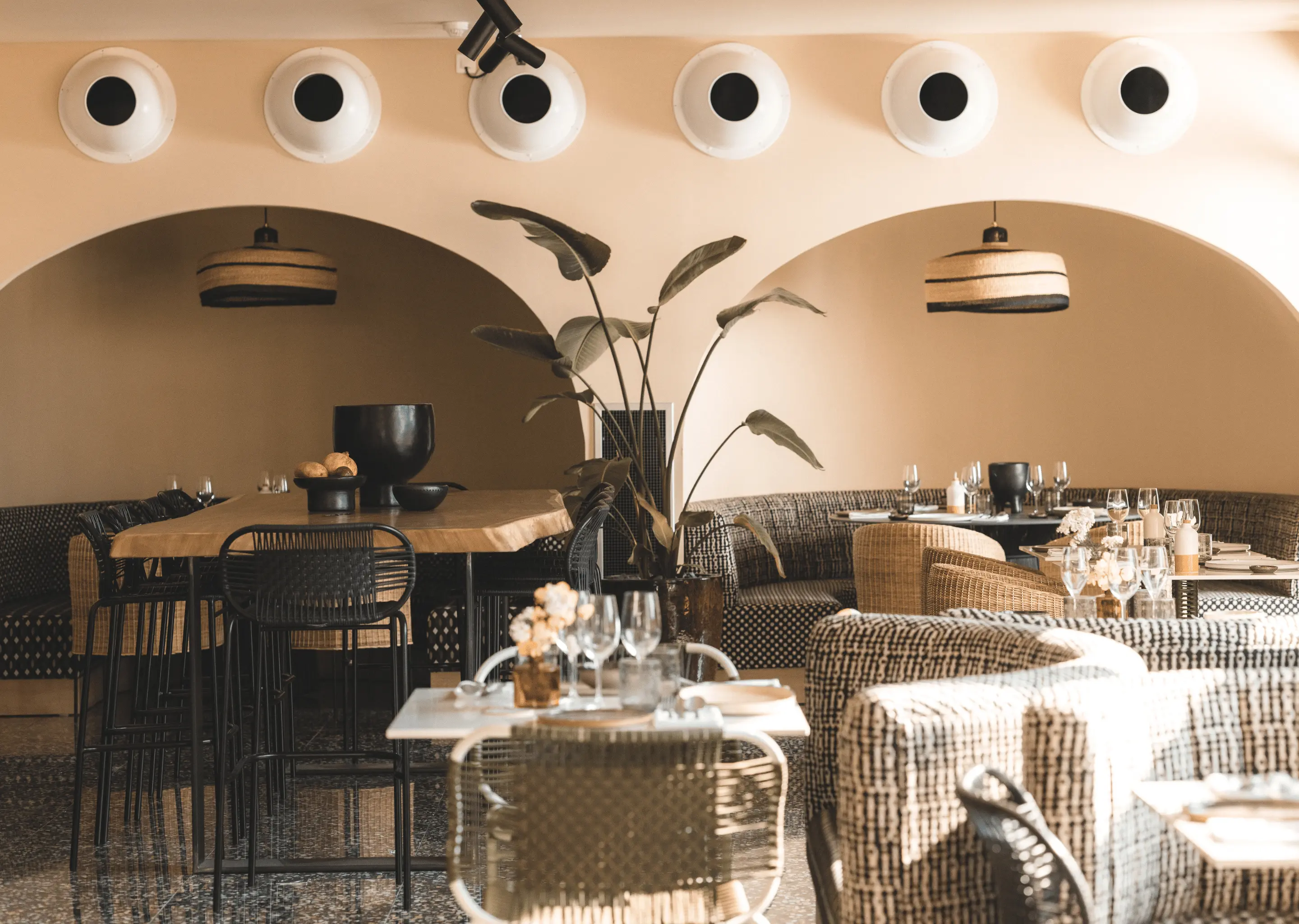 Interno accogliente del ristorante con posti a sedere curvi, tavoli in legno, sedie intrecciate e piante tropicali in vaso sotto archi beige.