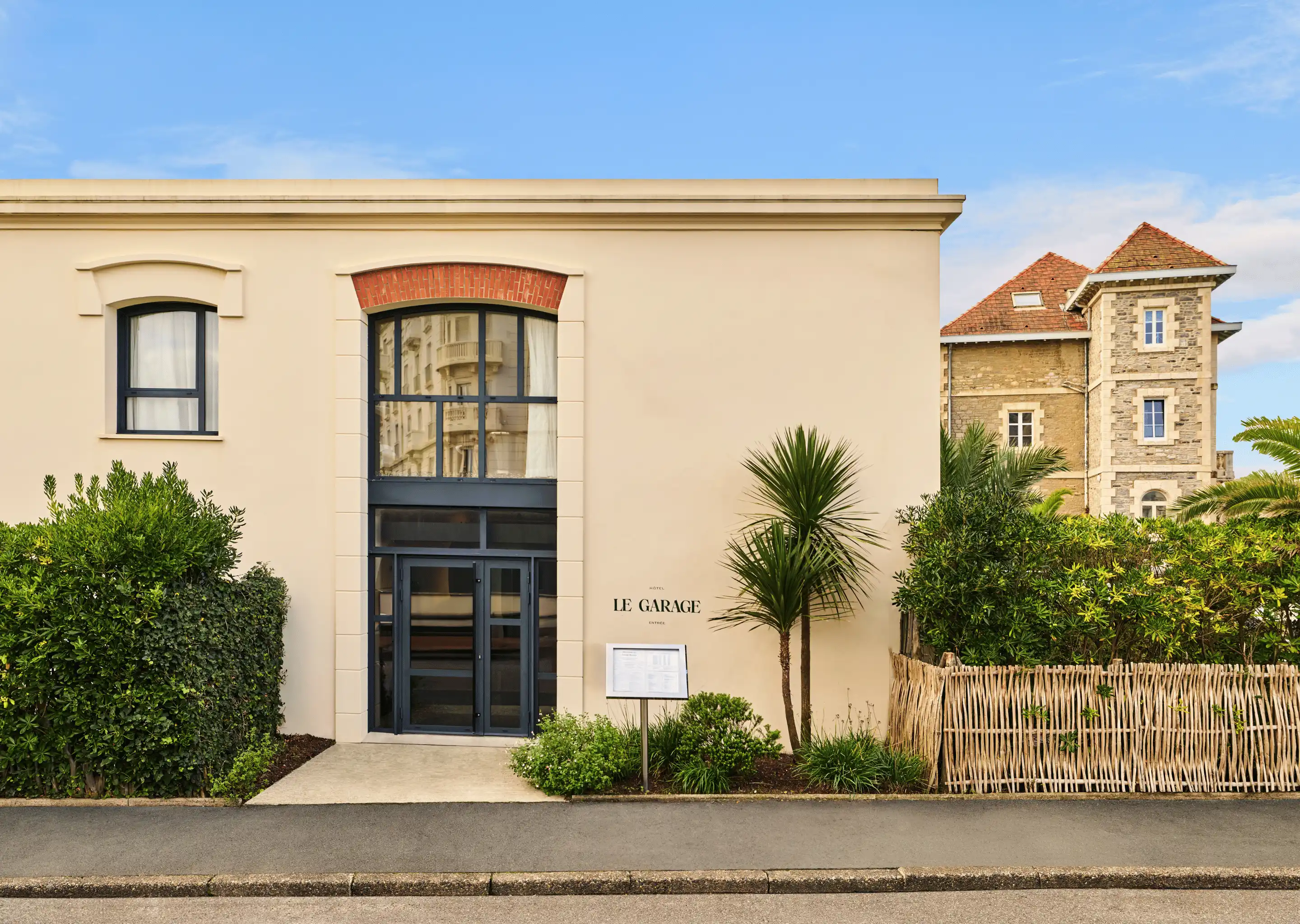 Facade of a building with tall arched glass doors and windows, labeled 'Hôtel Le Garage', surrounded by green shrubs and palm trees under a blue sky.