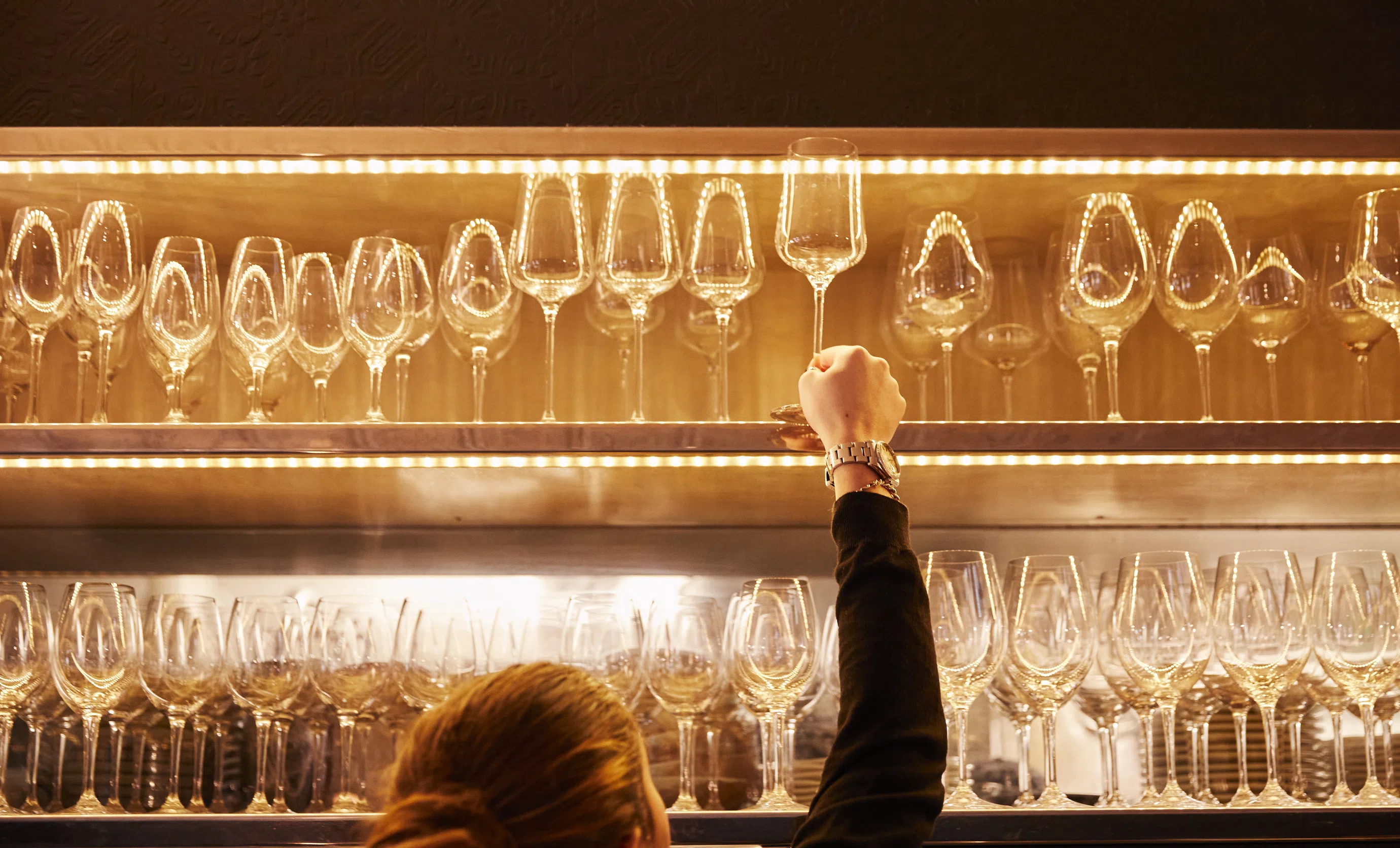 Close-up of a bartender tidying hanging wine glasses on an illuminated shelf, Experimental Group.