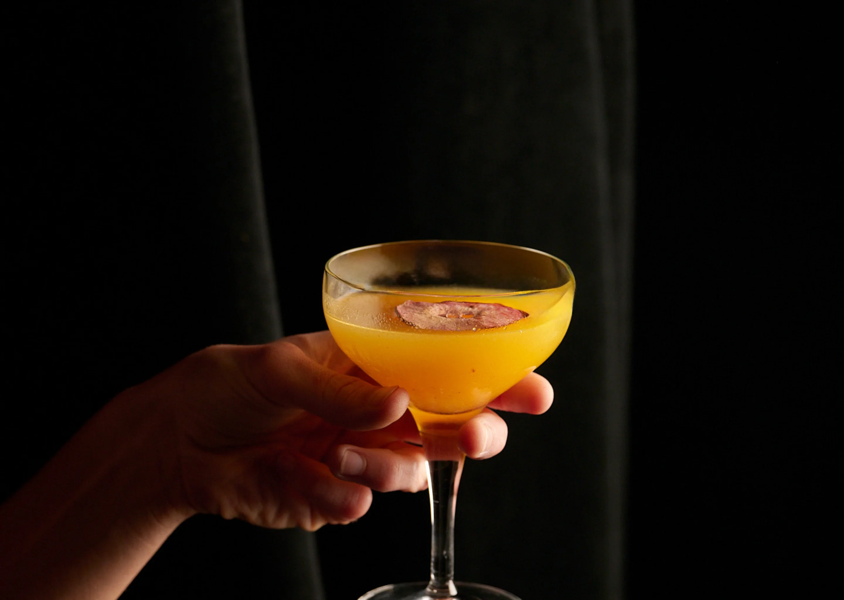 Close-up of a sophisticated yellow craft cocktail with a dried fruit garnish held at an Experimental Group bar.