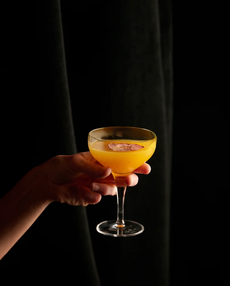Hand holding an elegant coupe glass filled with a golden yellow cocktail garnished with an apple slice at an Experimental Group bar.
image_generation_content_0 (Image de la table)