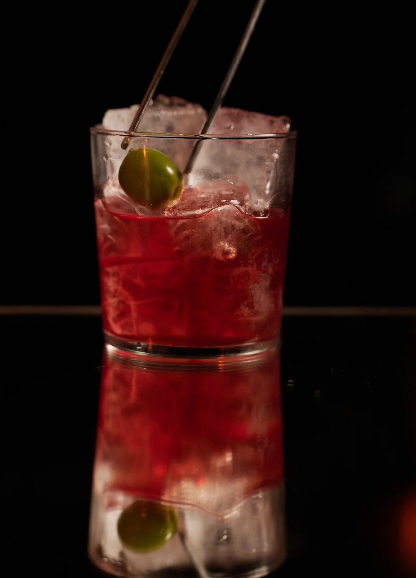 A bartender using tweezers to place a green olive garnish into a deep red cocktail at Experimental Cocktail Club.
