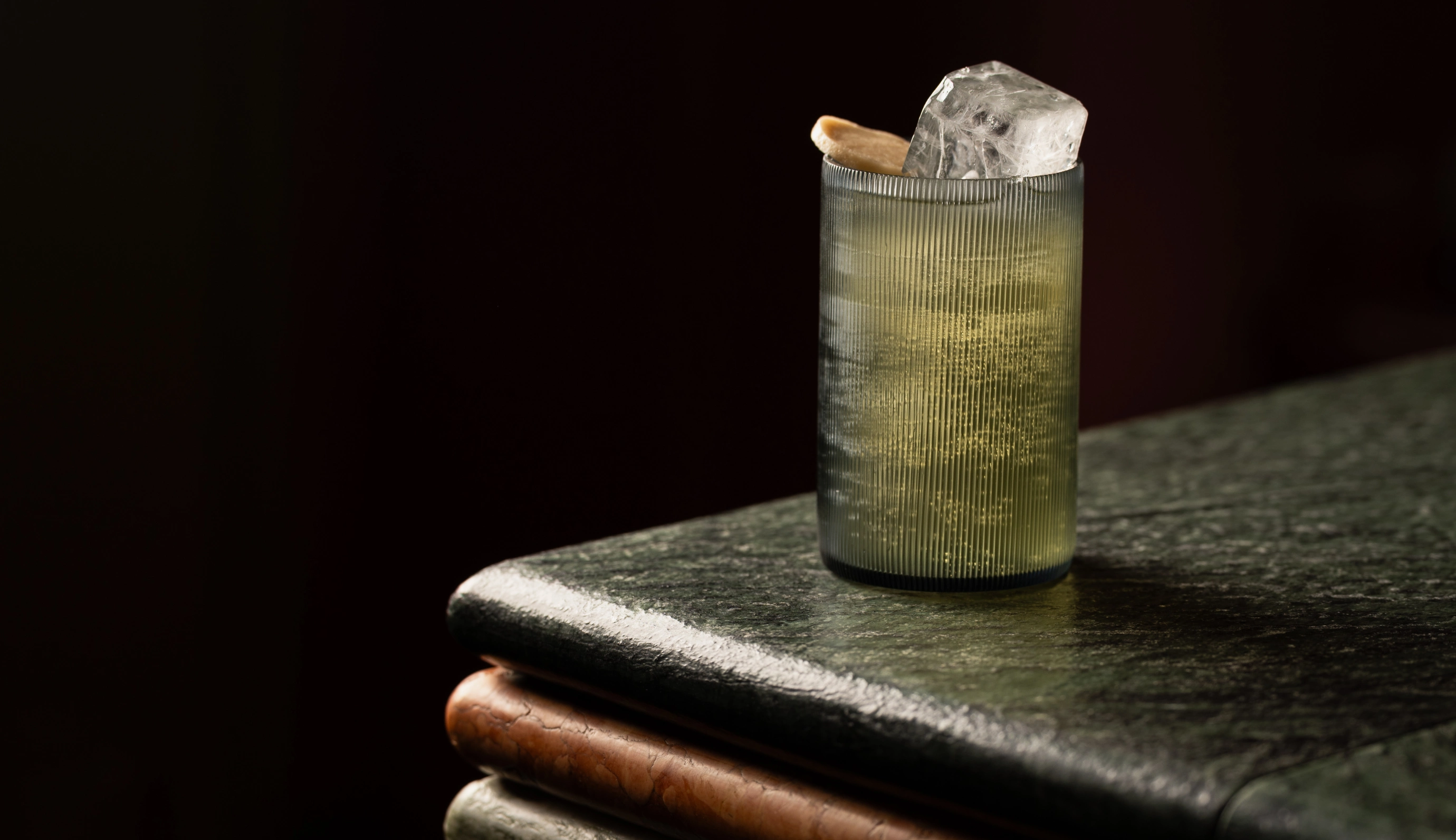 Close-up view of a signature cocktail in a textured glass on a dark stone table, intimate Experimental Group atmosphere.