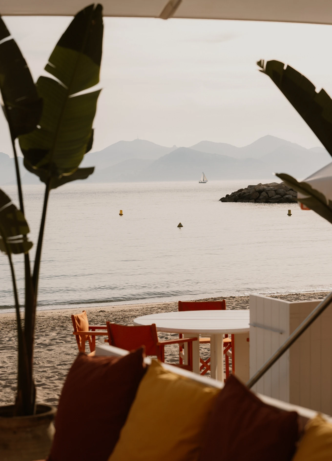 View of a calm bay with a sailboat in the distance, framed by tropical plants at an Experimental Group seaside property.