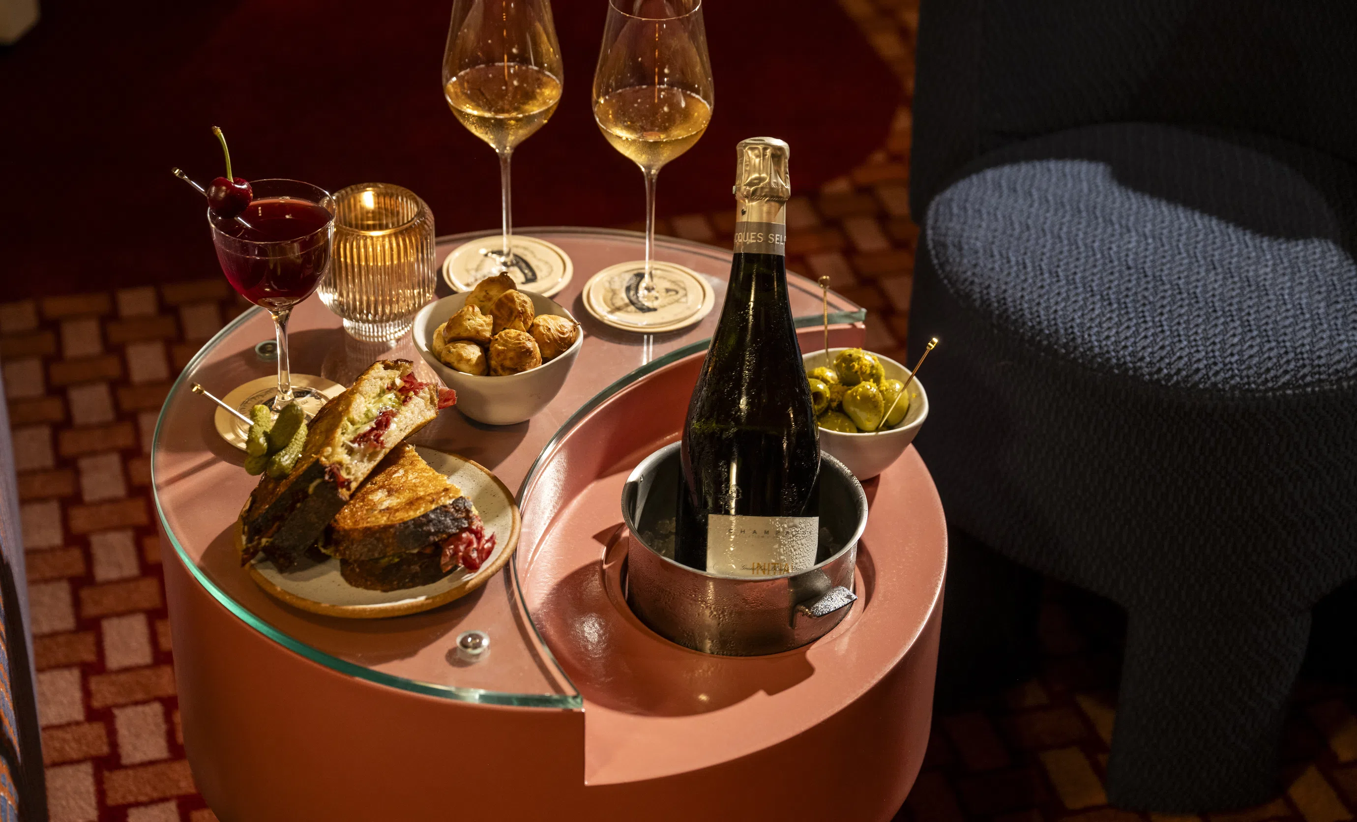 Mesa gourmet servida com uma garrafa de champanhe Jacques Selosse, lanches e um sanduíche em um lounge do Grupo Experimental.
