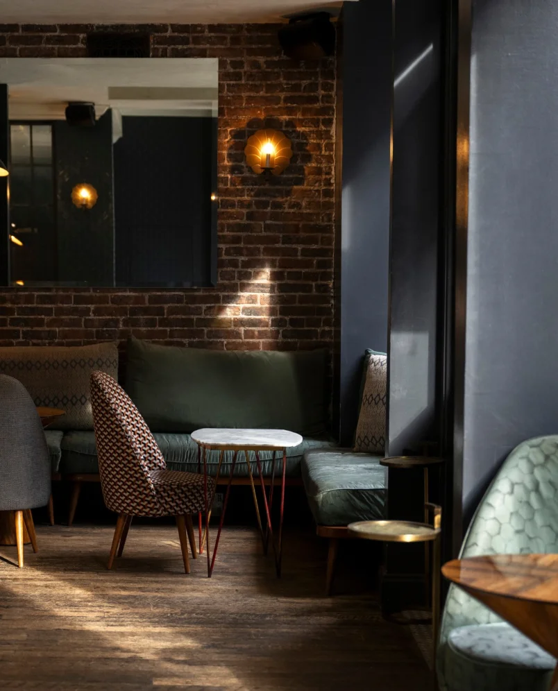 Intimate lounge corner with green velvet bench, marble table, patterned armchair, and brick wall at The Henrietta Hotel bar.