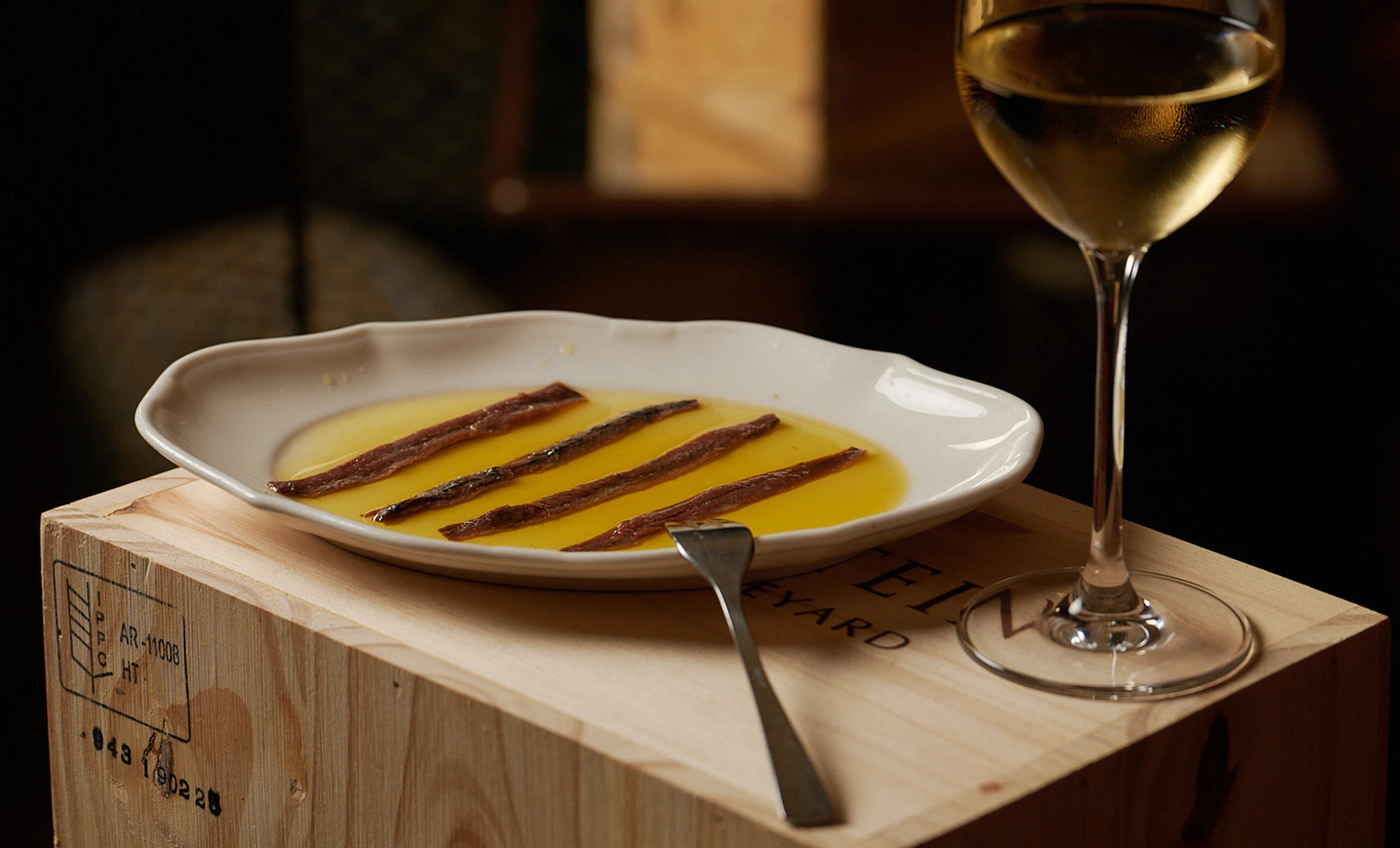 Un sofisticado plato de anchoas servido con aceite de oliva y una copa de vino blanco en una caja de vino de madera, parte de la experiencia culinaria de Regina Experimental, Biarritz.