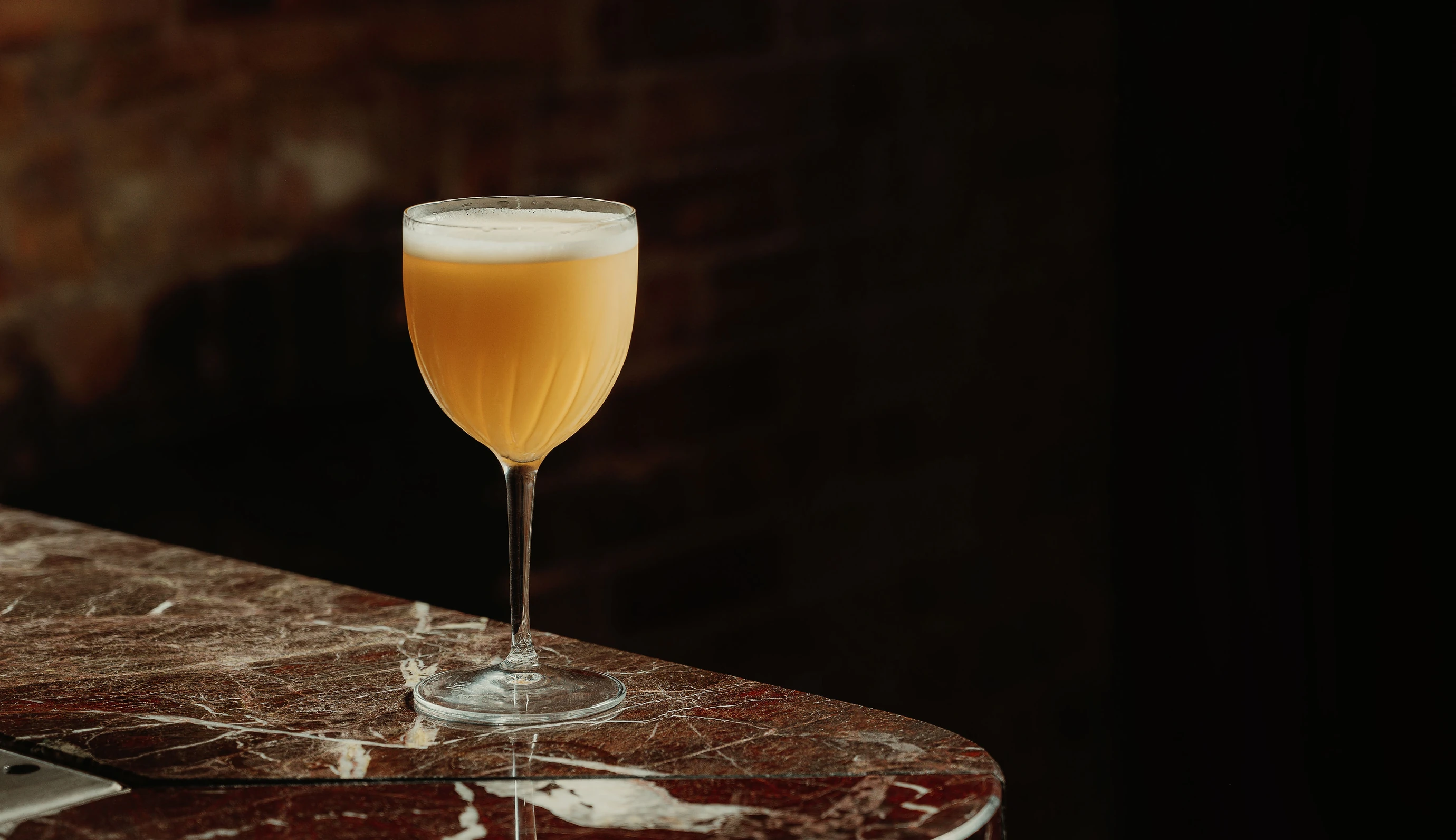 Gros plan d'un cocktail artisanal jaune emblématique servi dans une coupe en cristal sur une surface en marbre rouge poli dans une salle d'Experimental Group.