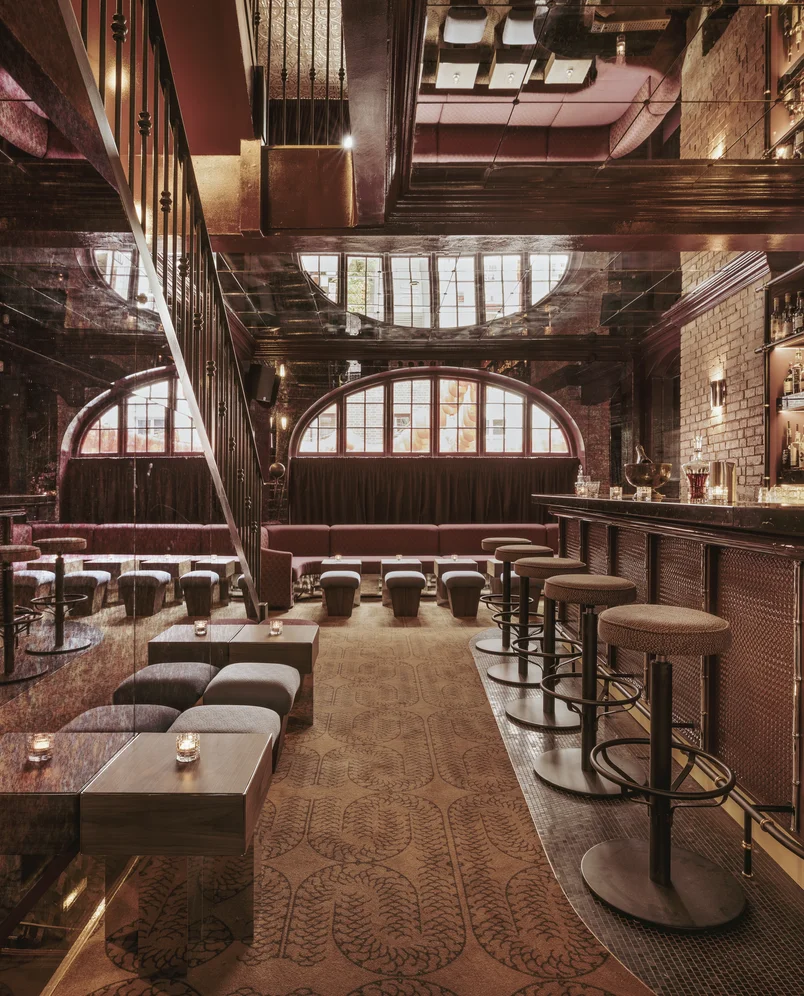 Vue intérieure d'un élégant bar à cocktails d'Experimental Group doté de fenêtres cintrées et de sièges en cuir.