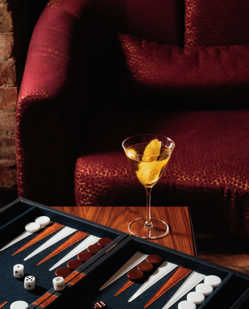 Cocktail raffiné dans un verre à pied à côté d'un jeu de backgammon sur une table, avec un canapé rouge en arrière-plan.