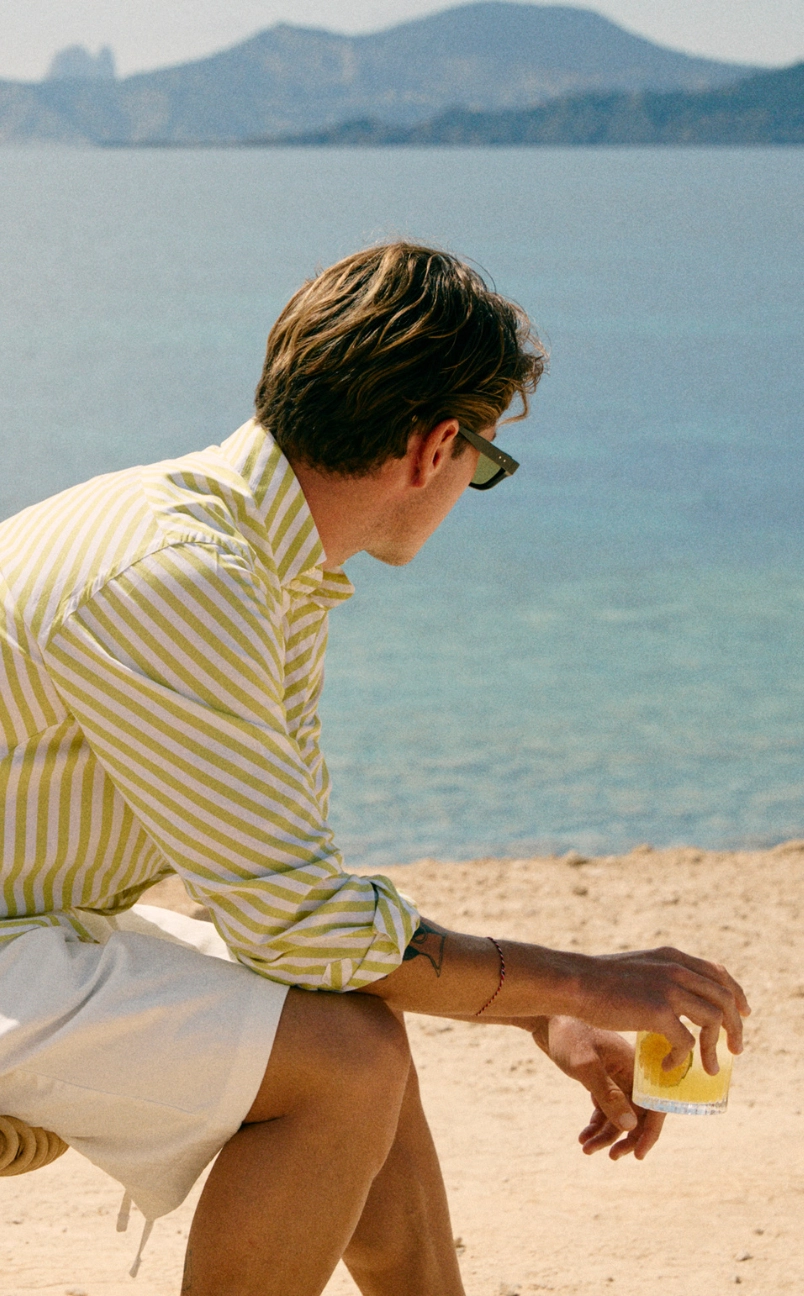 Man wearing a green and white striped shirt holding a cocktail on a sunny beach with the sea in the background.