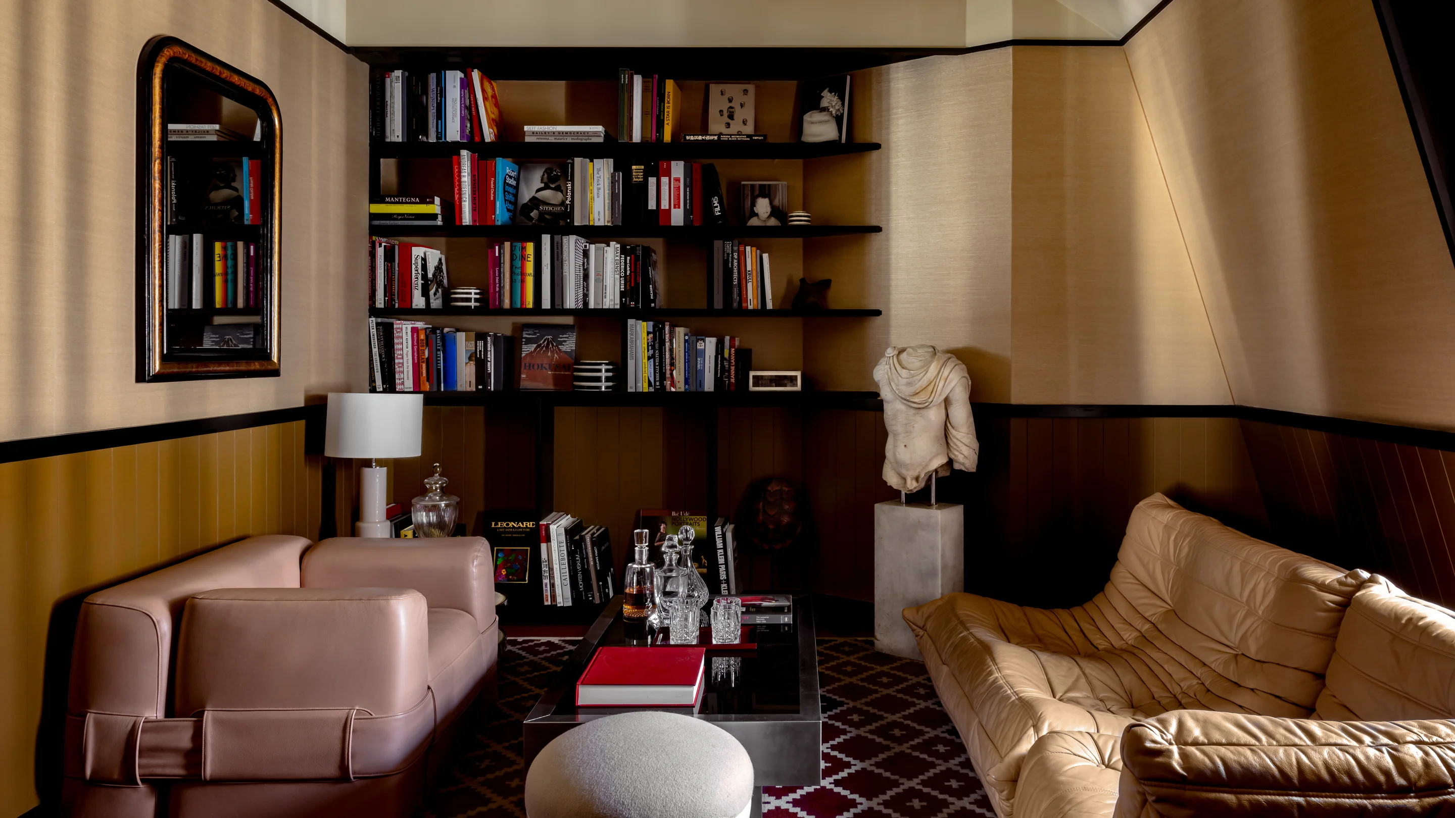 Salle de bibliothèque confortable avec fauteuil rose, canapé beige, étagère, torse de statue et carafes en verre sur la table.
