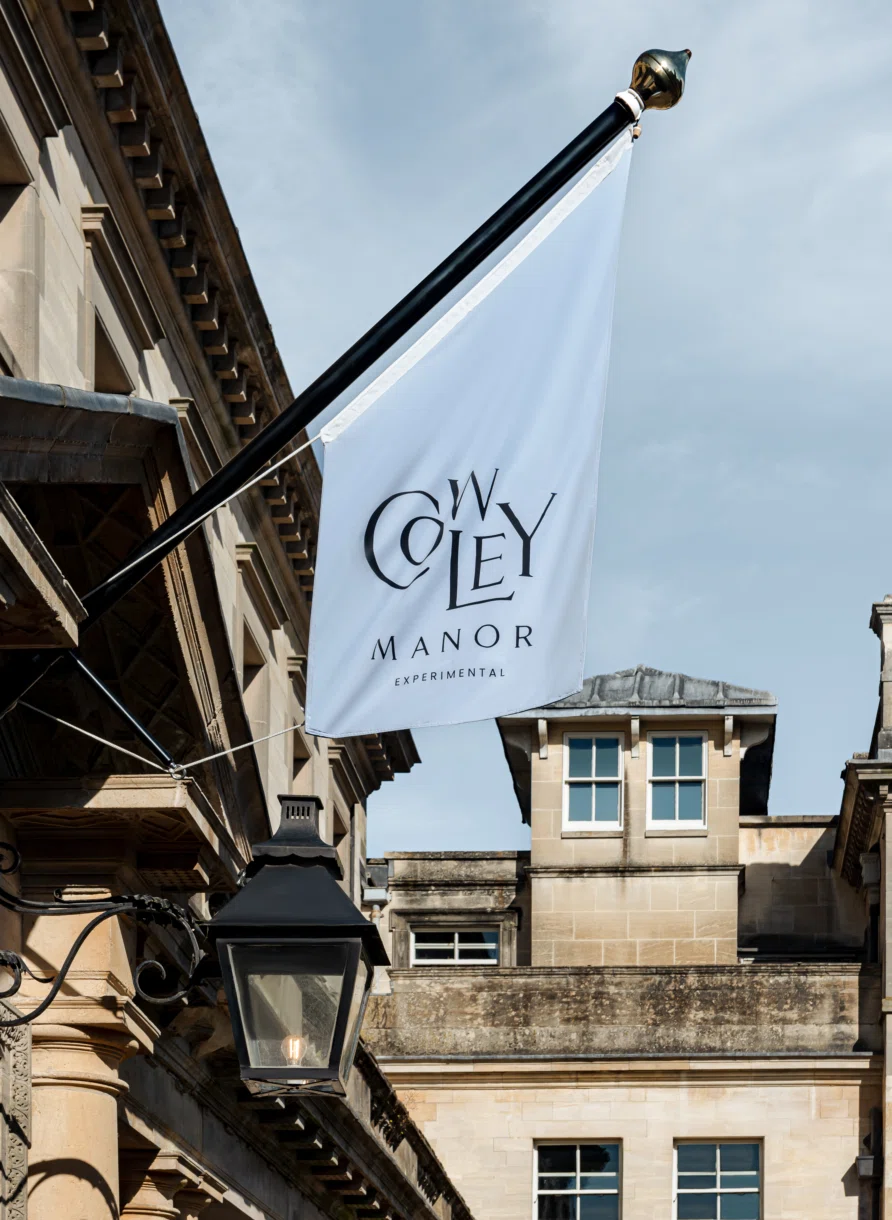 Bandiera bianca con il logo Cowley Manor Experimental appeso a un edificio in pietra beige accanto a una lanterna nera.