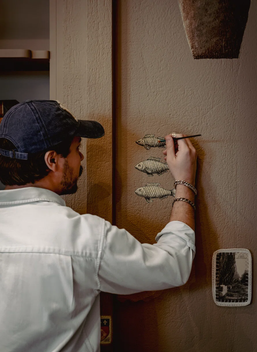 Hombre con gorra pintando tres dibujos de peces en una pared marrón texturizada.