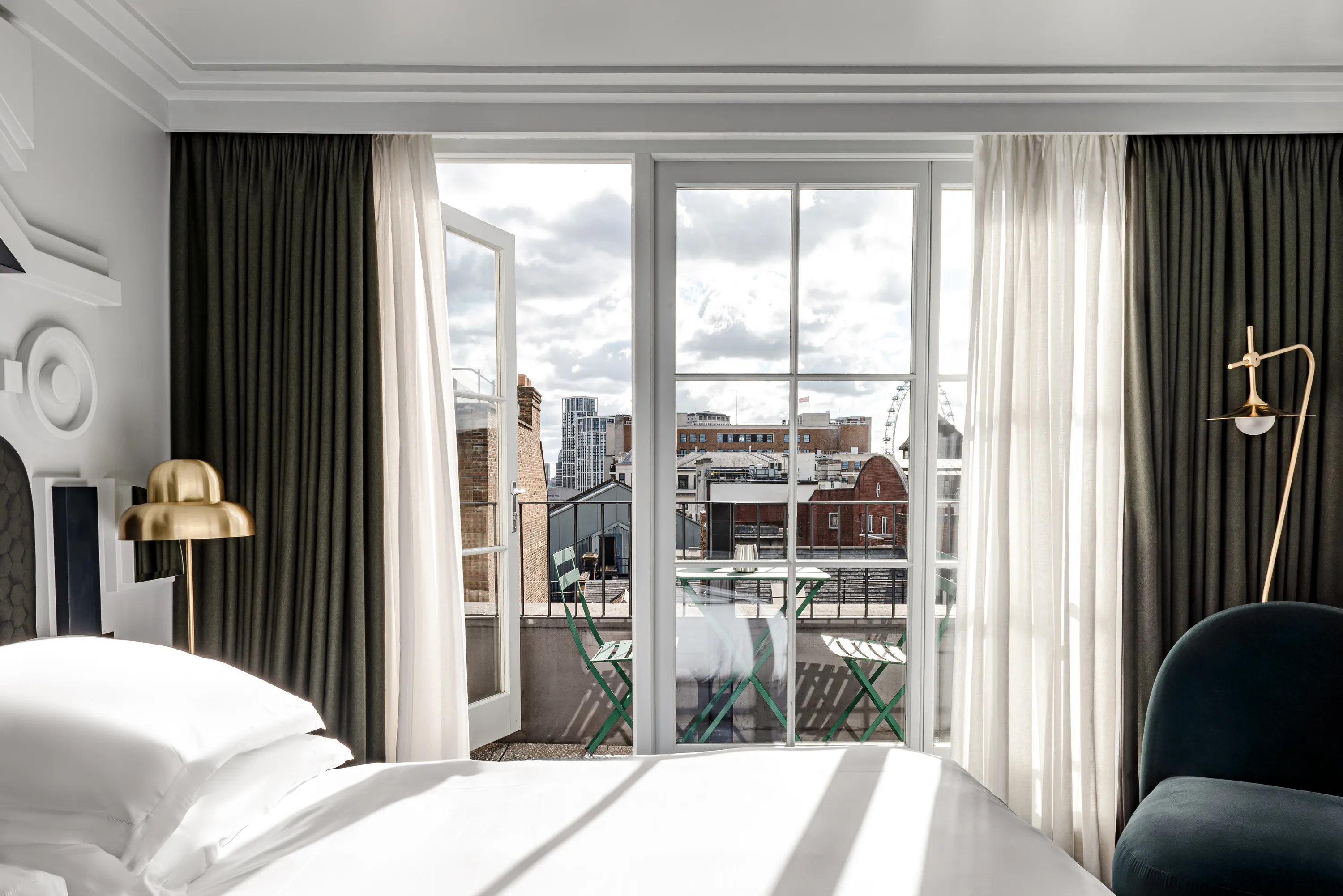 Camera d'albergo luminosa con letto bianco, lampade dorate, tende scure e balcone con vista sugli edifici della città.