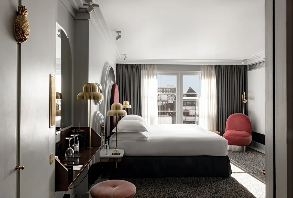 Moderna suite d'albergo con un grande letto, lampade dorate, una sedia rossa e ampie finestre con tende.