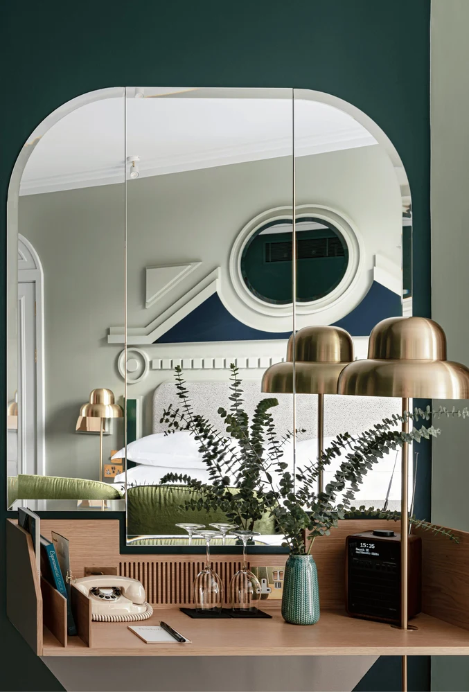 Desk with vintage phone, lamp, vase with greenery, and mirrored wall reflecting a bed and decor.