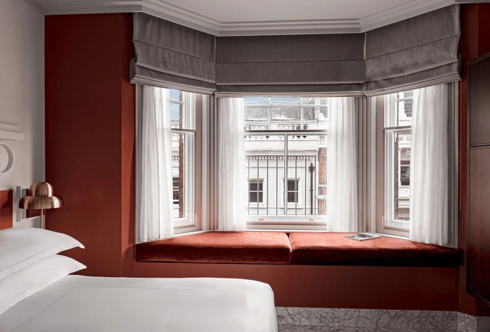 Suite d'albergo con letto bianco, sedile vicino al finestrino color ruggine e ampie vetrate con tende trasparenti.