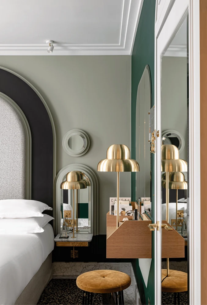 Moderno angolo della camera d'albergo con lampada dorata, sgabello rotondo, specchi ad arco e parte di un letto bianco.