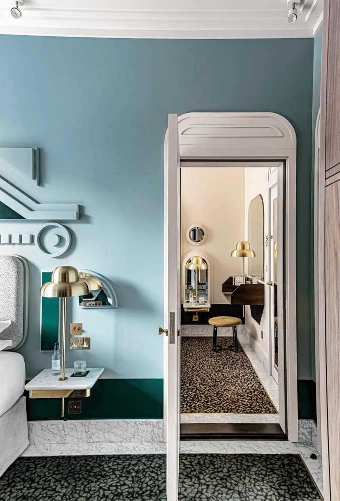 Porta aperta che mostra una camera da letto di una suite d'albergo con una parete blu, una lampada dorata e una zona vanità all'interno.