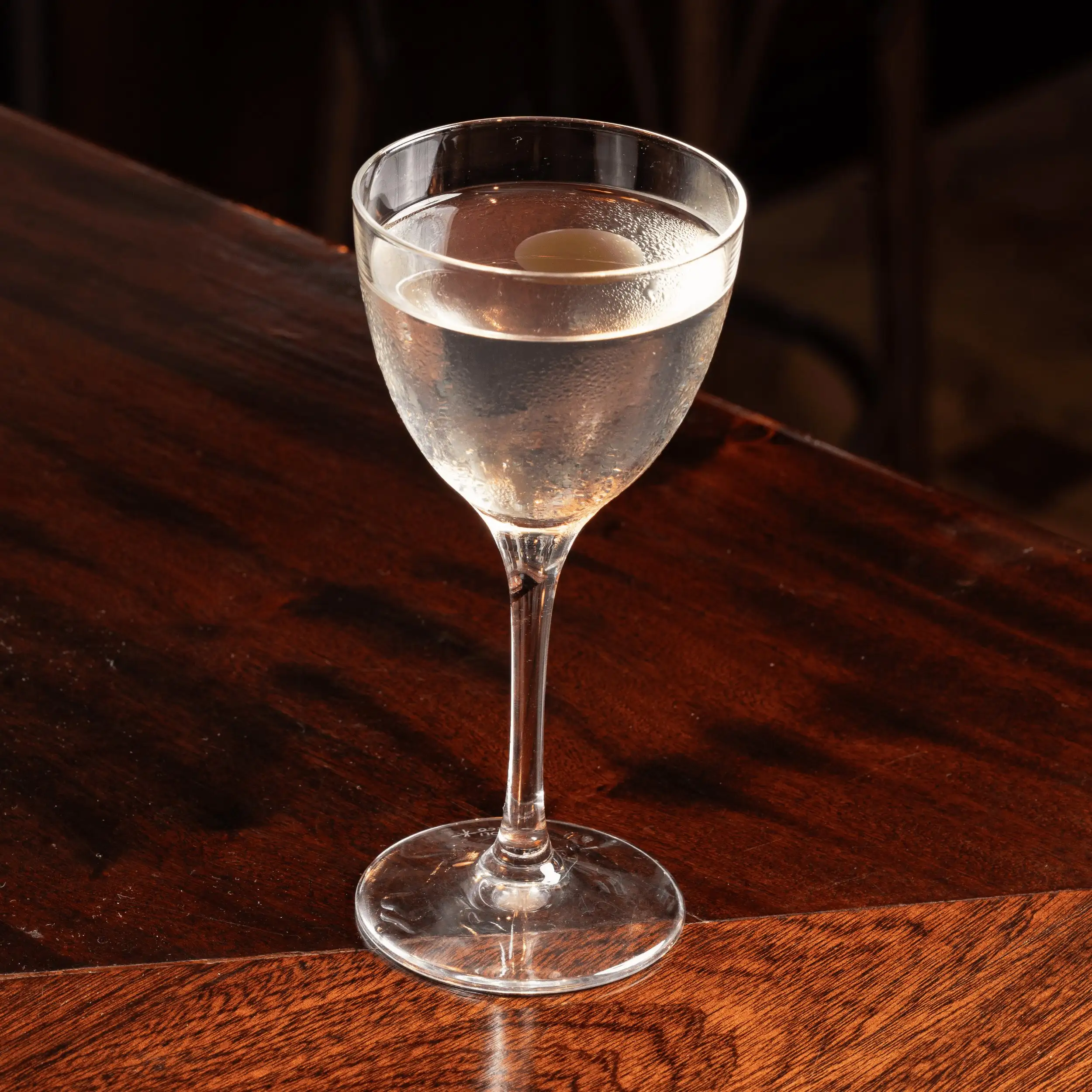 Bicchiere da cocktail trasparente con una bevanda fresca e una guarnizione rotonda su una superficie di legno scuro.