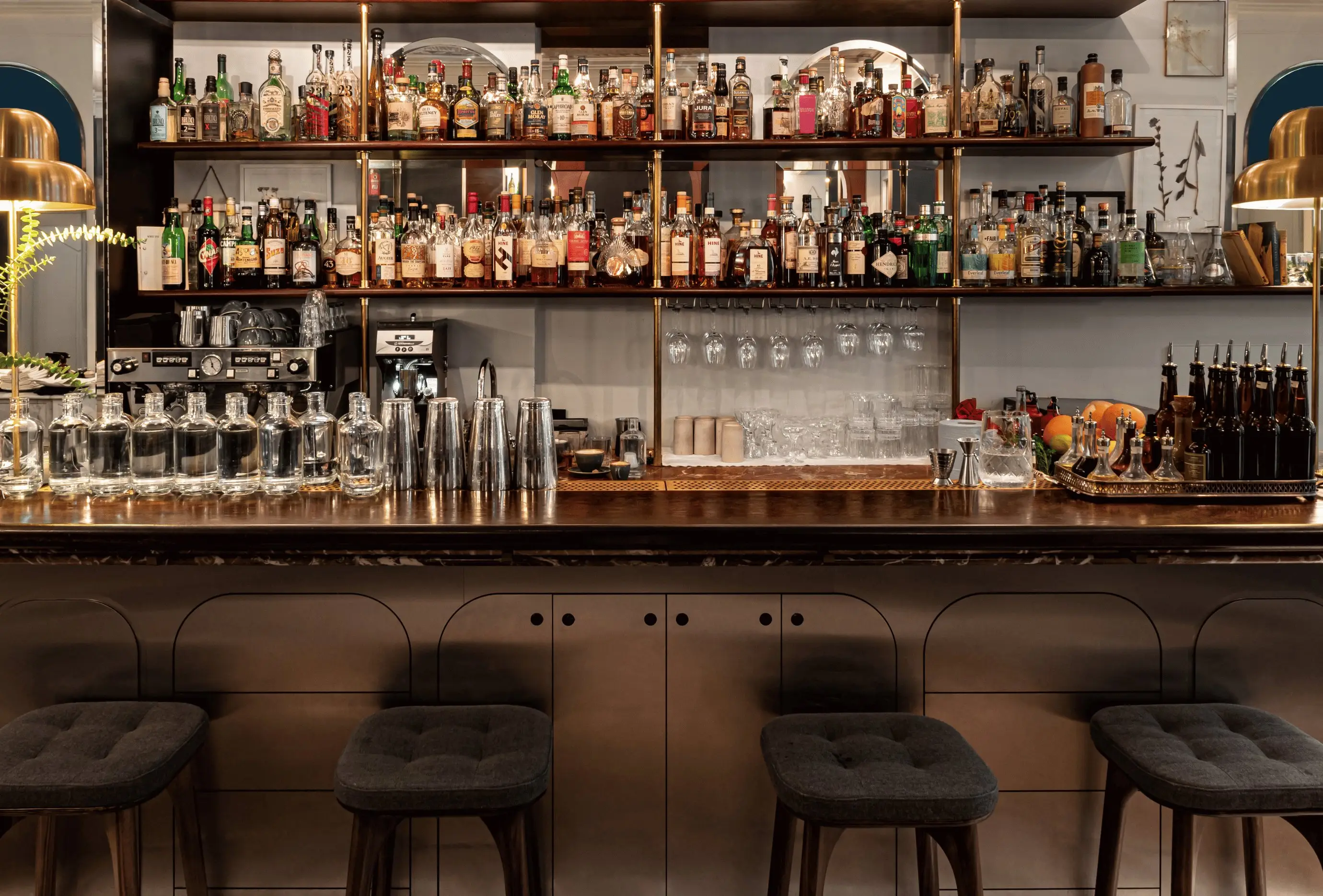 Bancone da bar con quattro sgabelli, bottiglie di vetro, shaker per cocktail e bottiglie di liquore su scaffali con illuminazione calda.