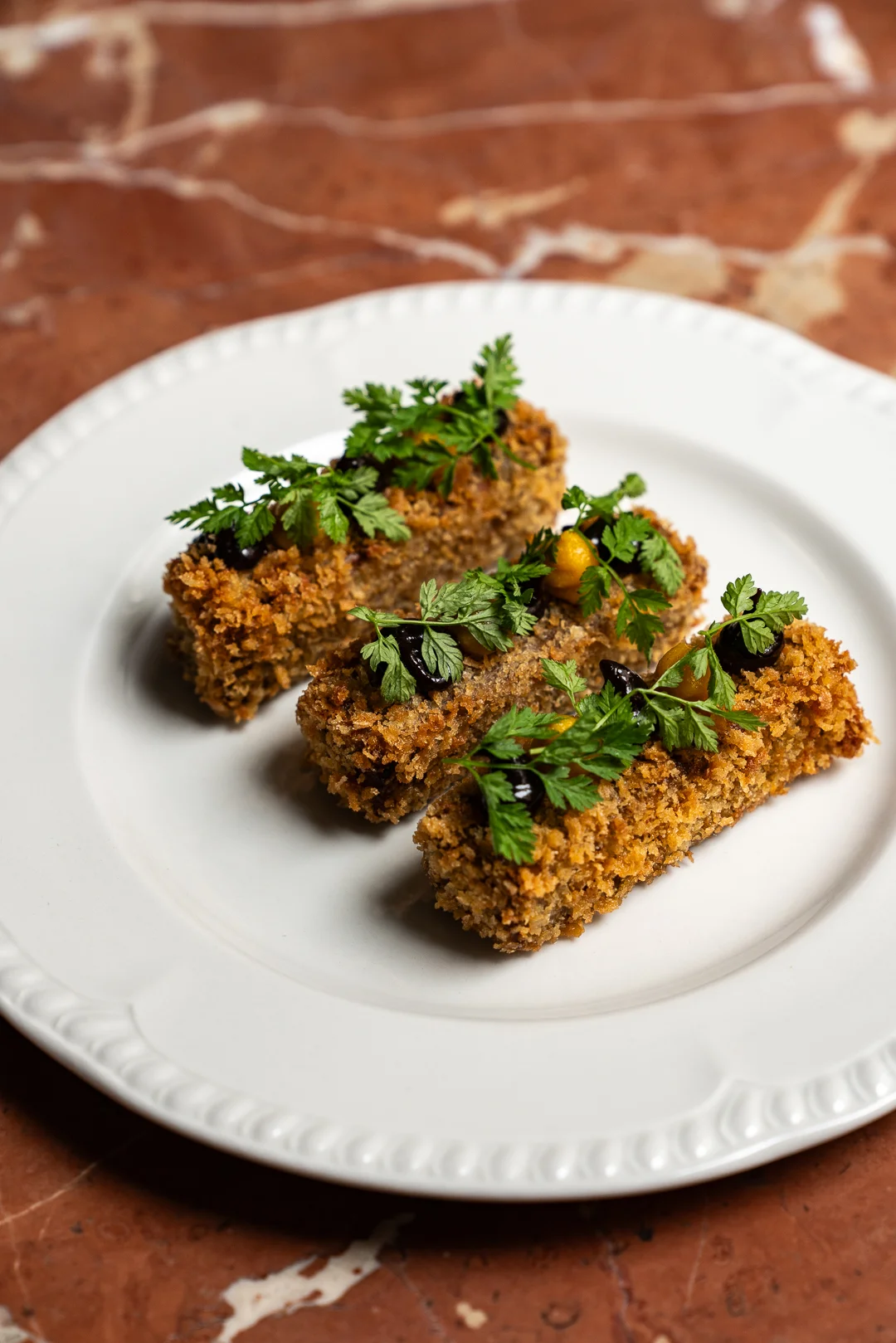 Tre crocchette rettangolari fritte croccanti guarnite con erbe verdi su un piatto bianco.