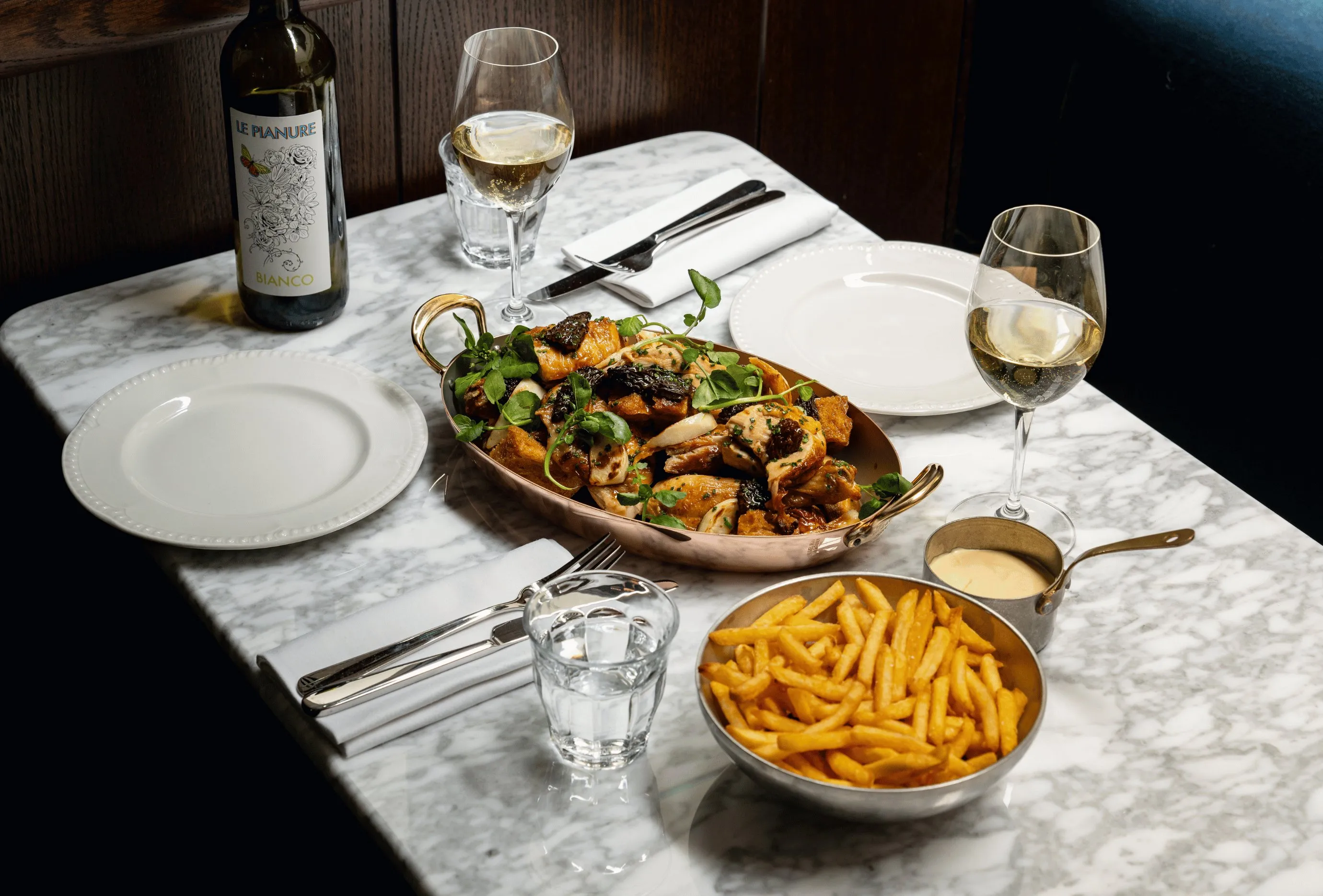 Servizio da tavola con pollo arrosto, patatine fritte con salsa, due bicchieri di vino bianco e una bottiglia di vino su marmo.