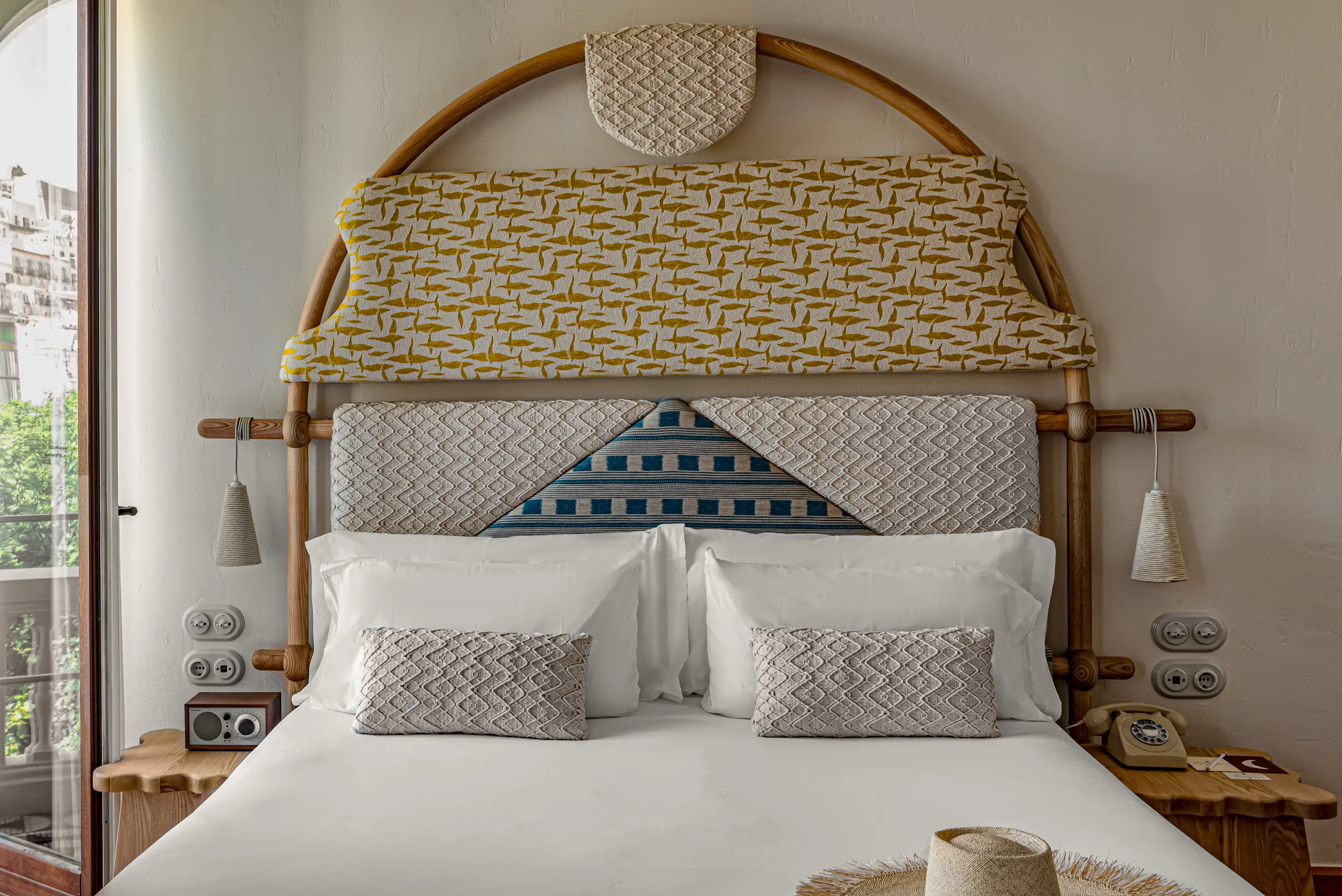 Letto d'albergo con testiera a motivi geometrici, cuscini bianchi, tavolini in legno e telefono rotativo vintage.