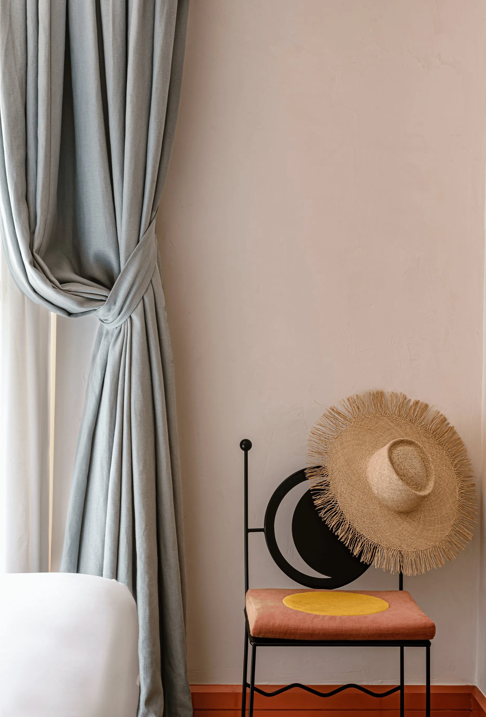 Sedia in metallo nero con un cappello di paglia sullo schienale accanto a una tenda grigia annodata e un letto bianco.