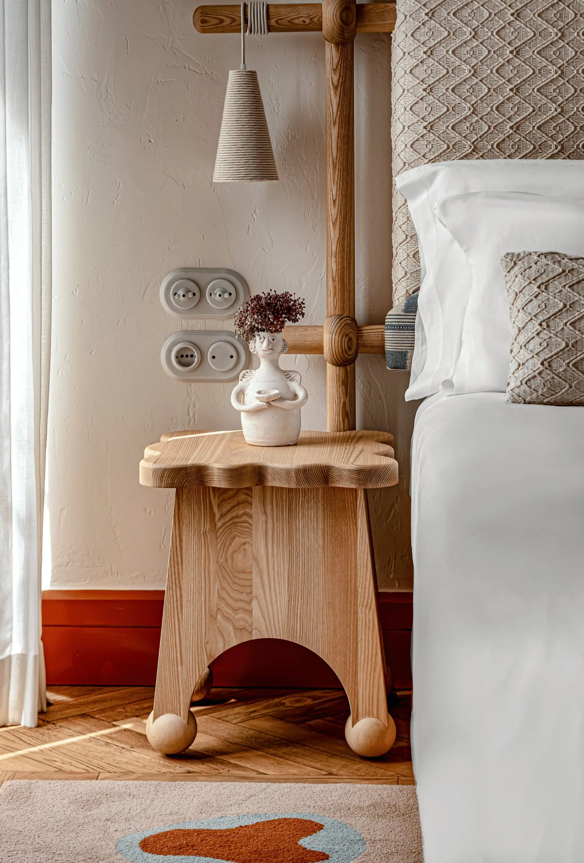 Wooden bedside table with a vase holding dried flowers next to a bed with textured cushions.