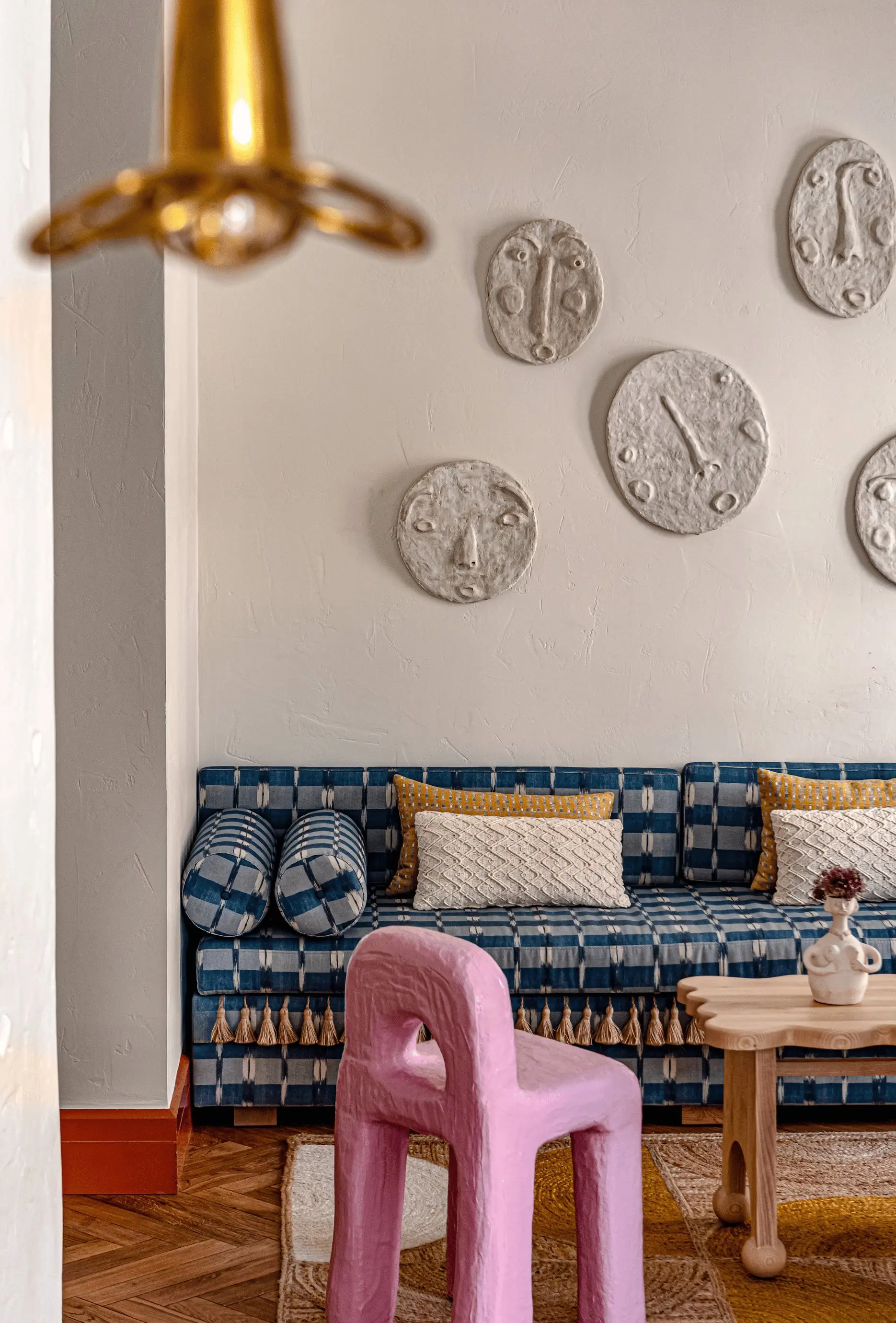 Divano a quadretti blu con cuscini e nappine, sedia scultorea rosa, tavolo in legno e placche decorative da parete.