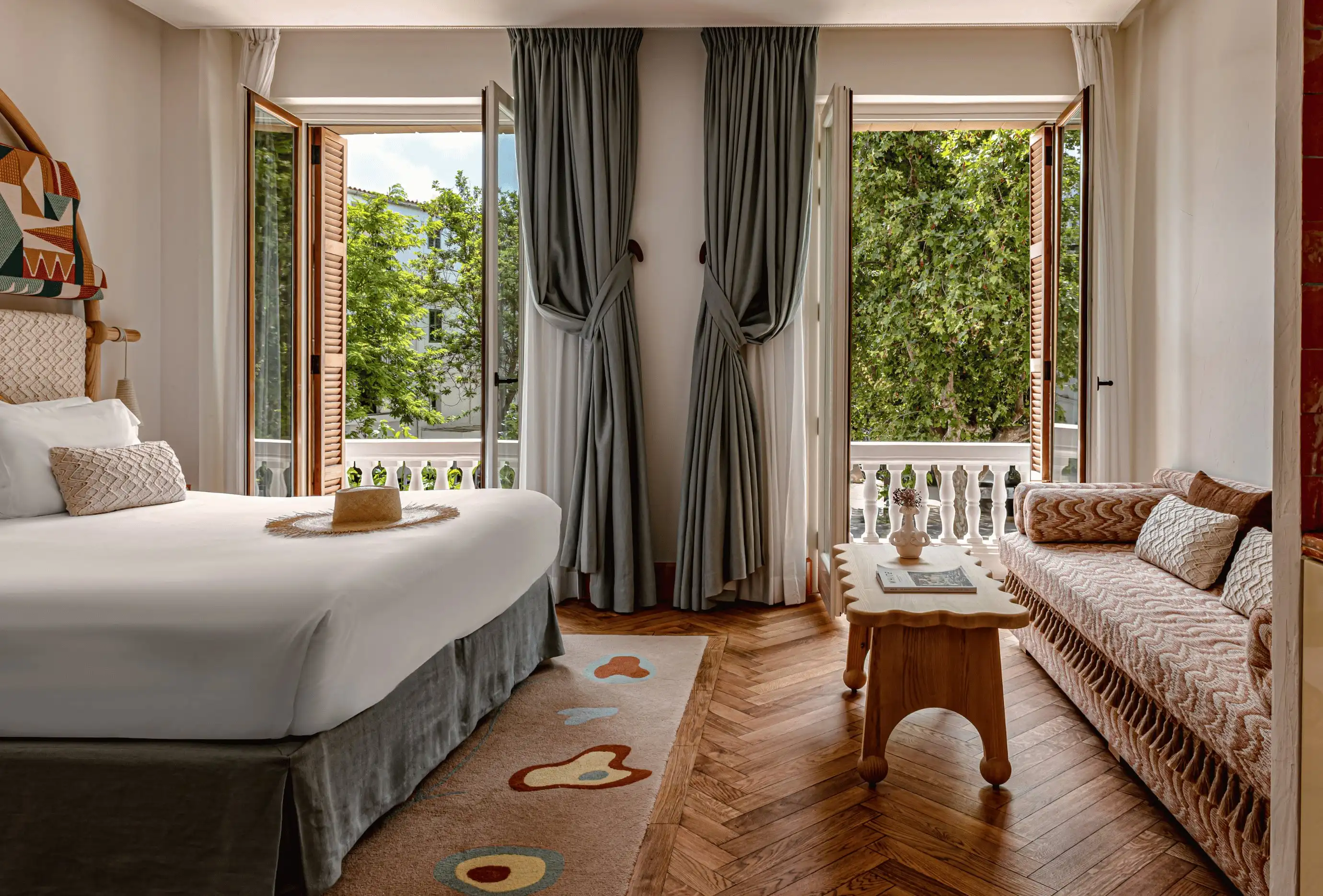 Camera d'albergo con letto, divano a motivi geometrici, tavolo in legno e finestre aperte che mostrano alberi verdi all'esterno.