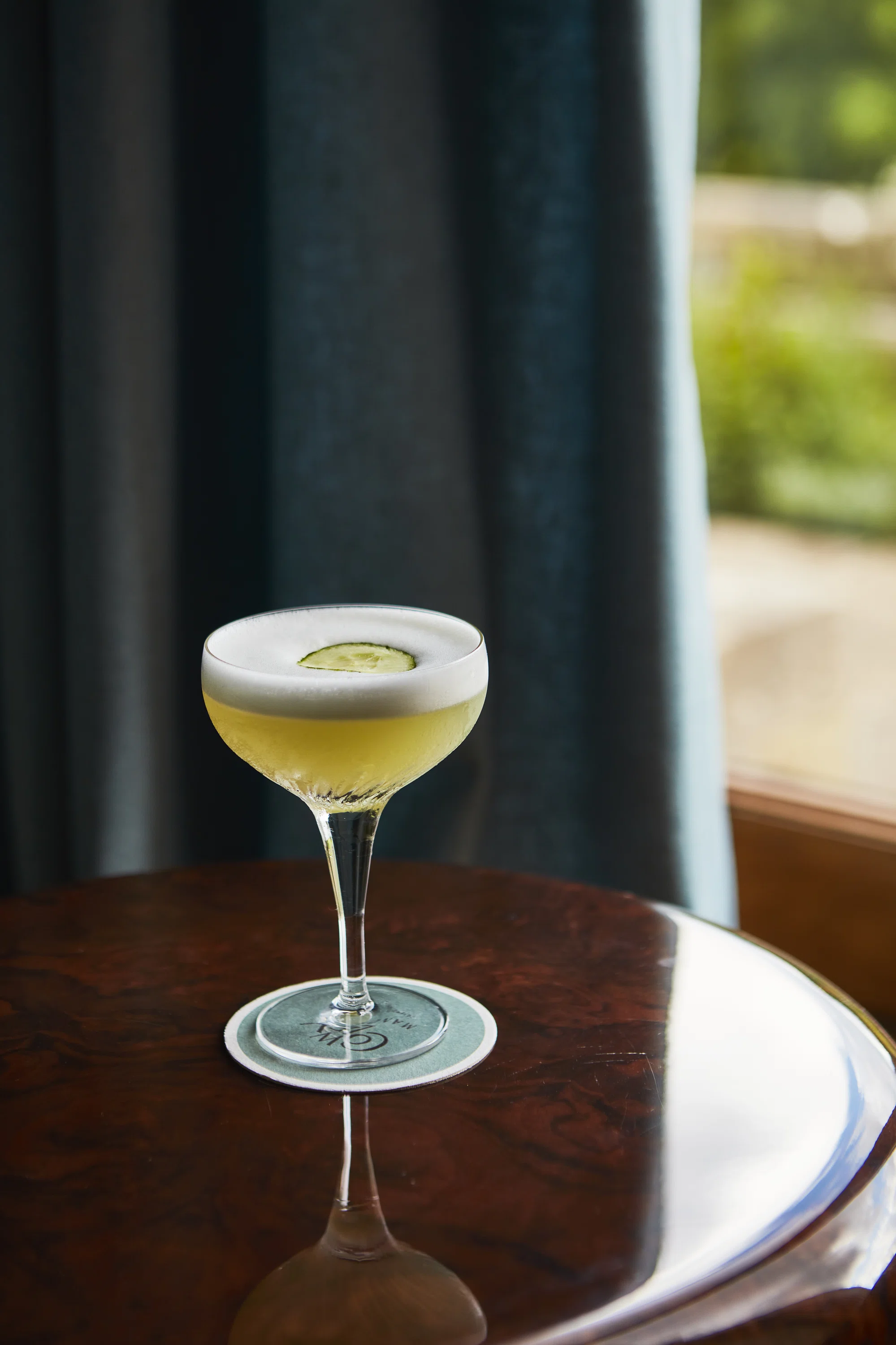 Cocktail glass with a yellow drink and foam topped with a cucumber slice on a coaster and wooden table.