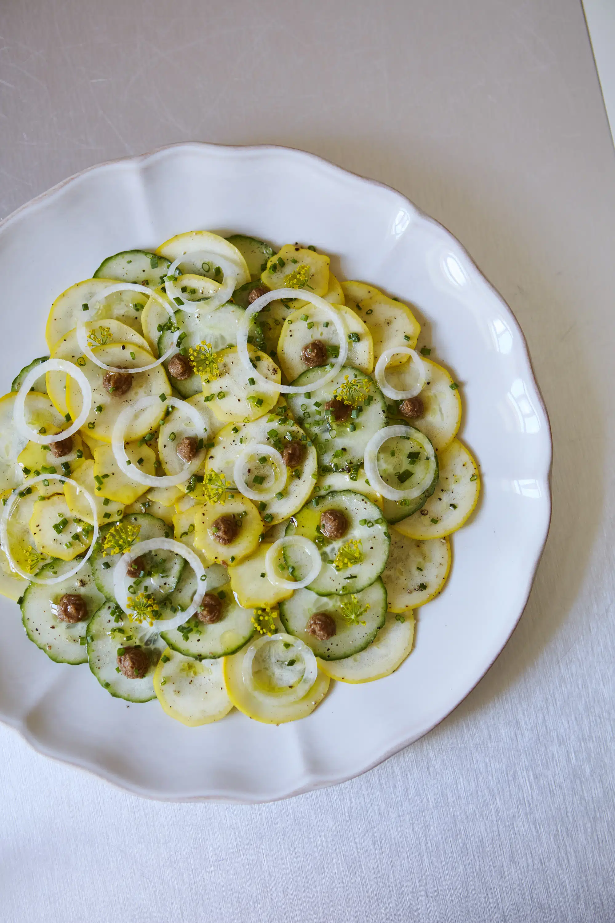 Piatto bianco con cetriolo a fette, zucca gialla, anelli di cipolla, erba cipollina e piccole cucchiaiate di crema marrone.