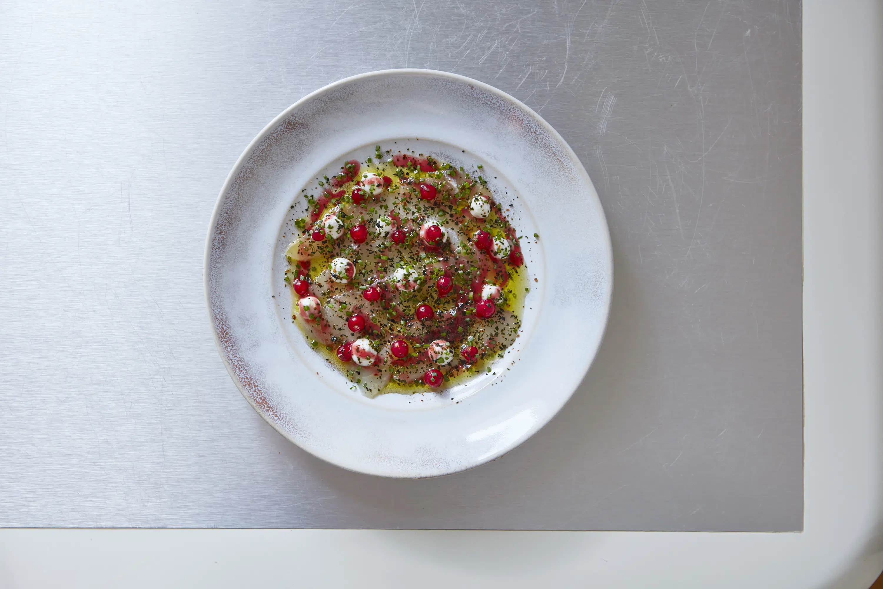 Piatto bianco con pesce tagliato a fettine sottili, frutti rossi, puntini cremosi, erbe aromatiche e olio su una superficie metallica.