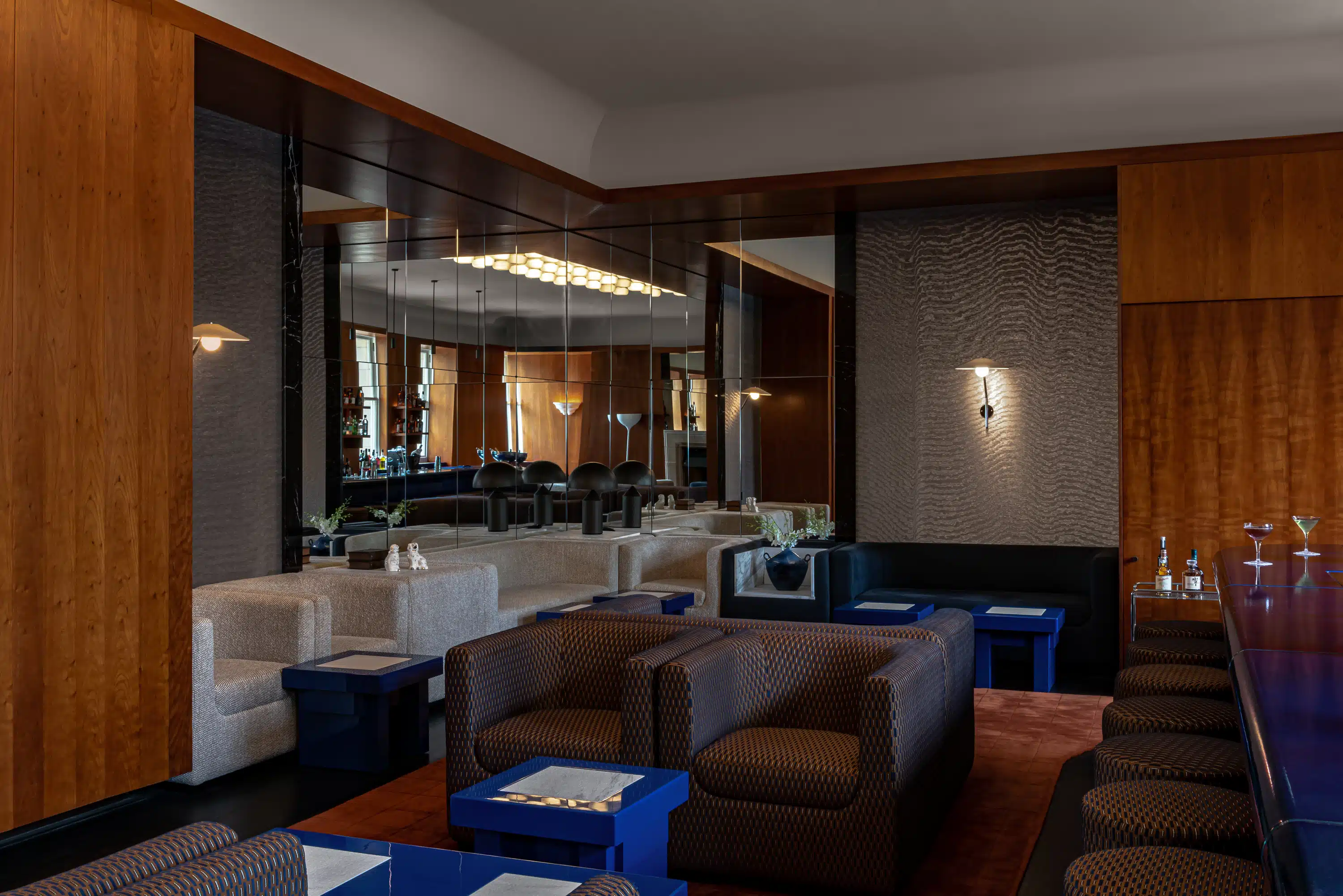 Moderno bar dell'hotel con poltrone a motivi geometrici, tavoli blu, parete a specchio e una fila di sgabelli da bar con bevande.