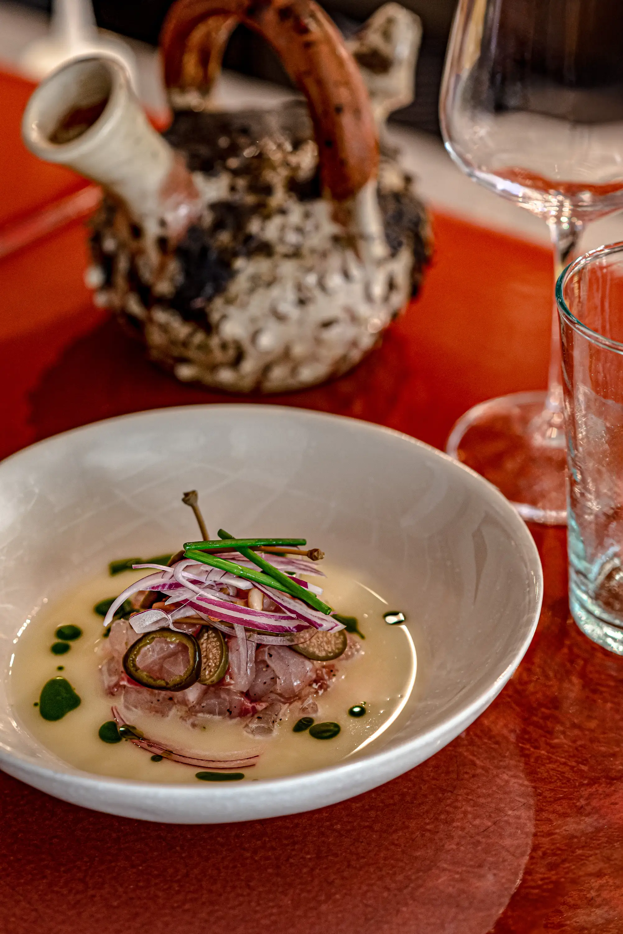 Ciotola bianca con ceviche di pesce crudo condito con cipolla rossa, capperi e contorno verde sul tavolo rosso.