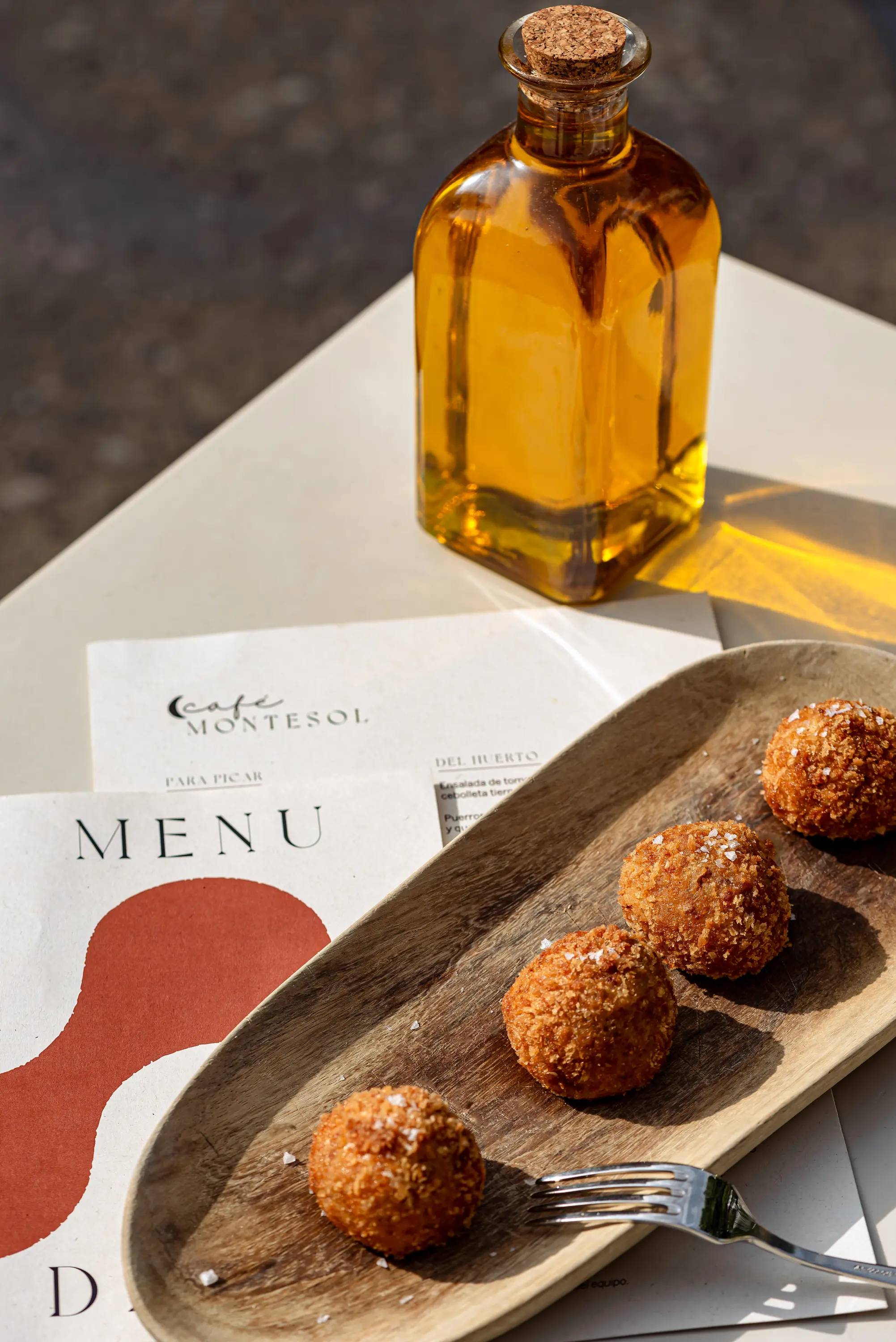 Quattro polpette fritte cosparse di sale su un piatto di legno accanto a una forchetta e una bottiglia nei menu.