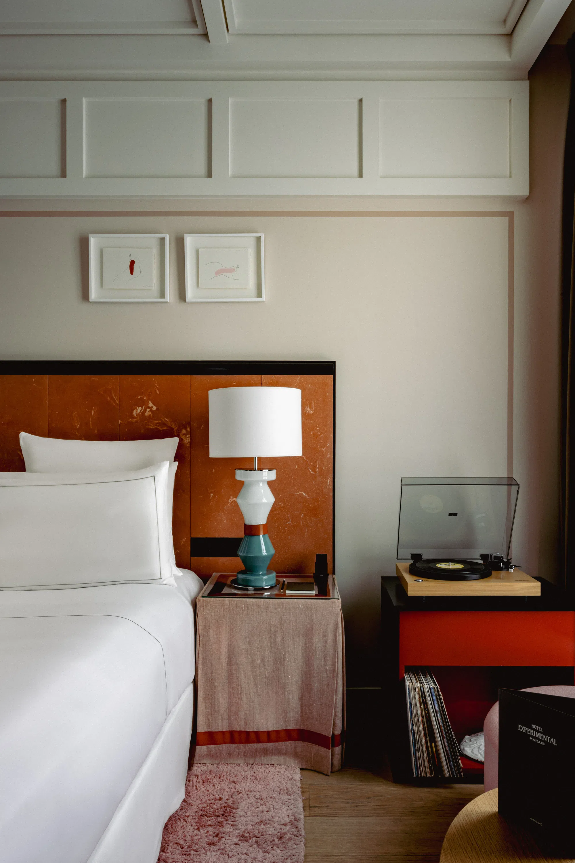 Habitación de hotel con cama blanca, lámpara colorida en la mesita de noche, arte enmarcado y tocadiscos en un armario rojo.