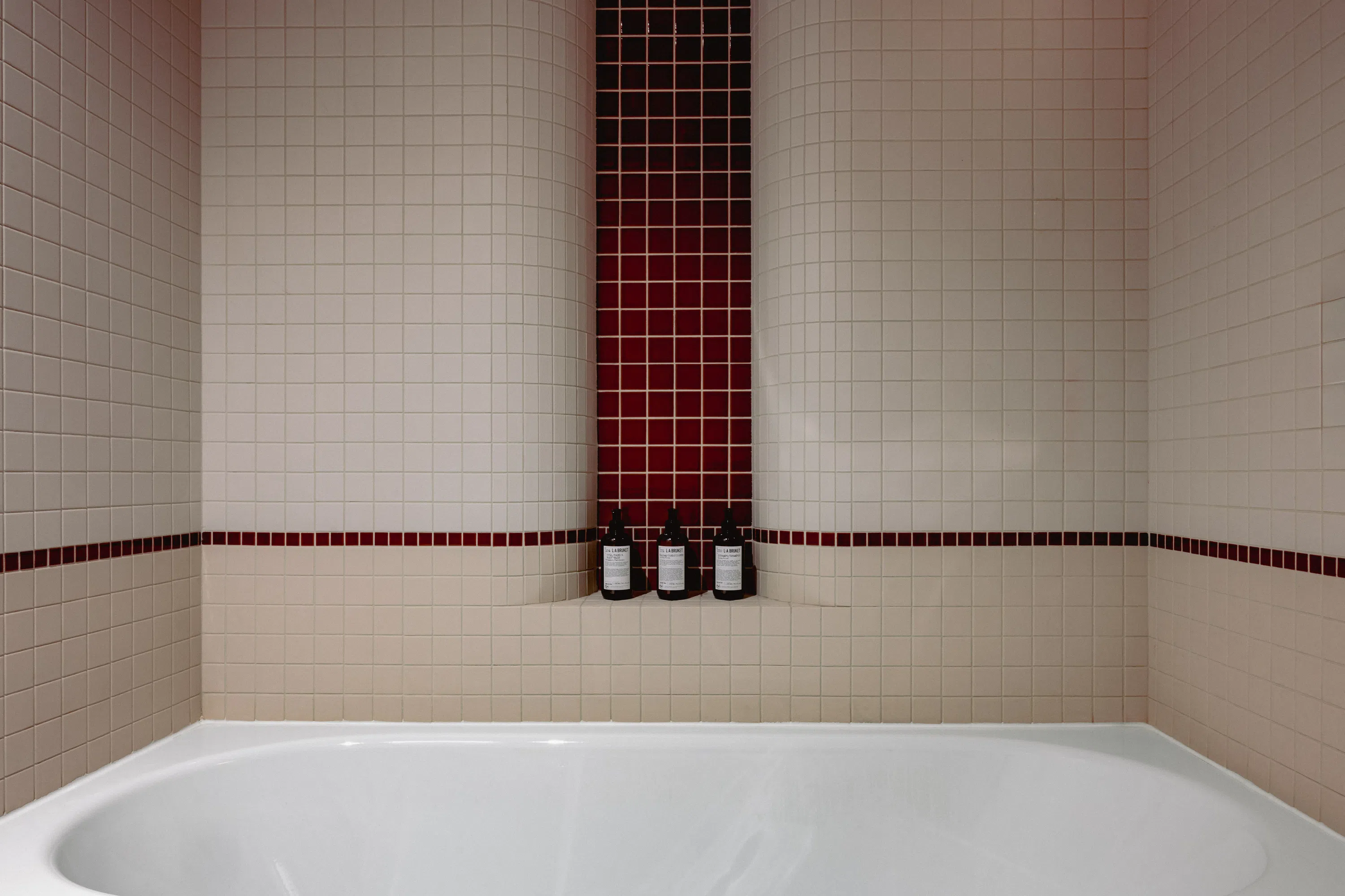 Bañera blanca con paredes de azulejos de color beige y una sección vertical de azulejos de color rojo oscuro con capacidad para tres botellas negras.
